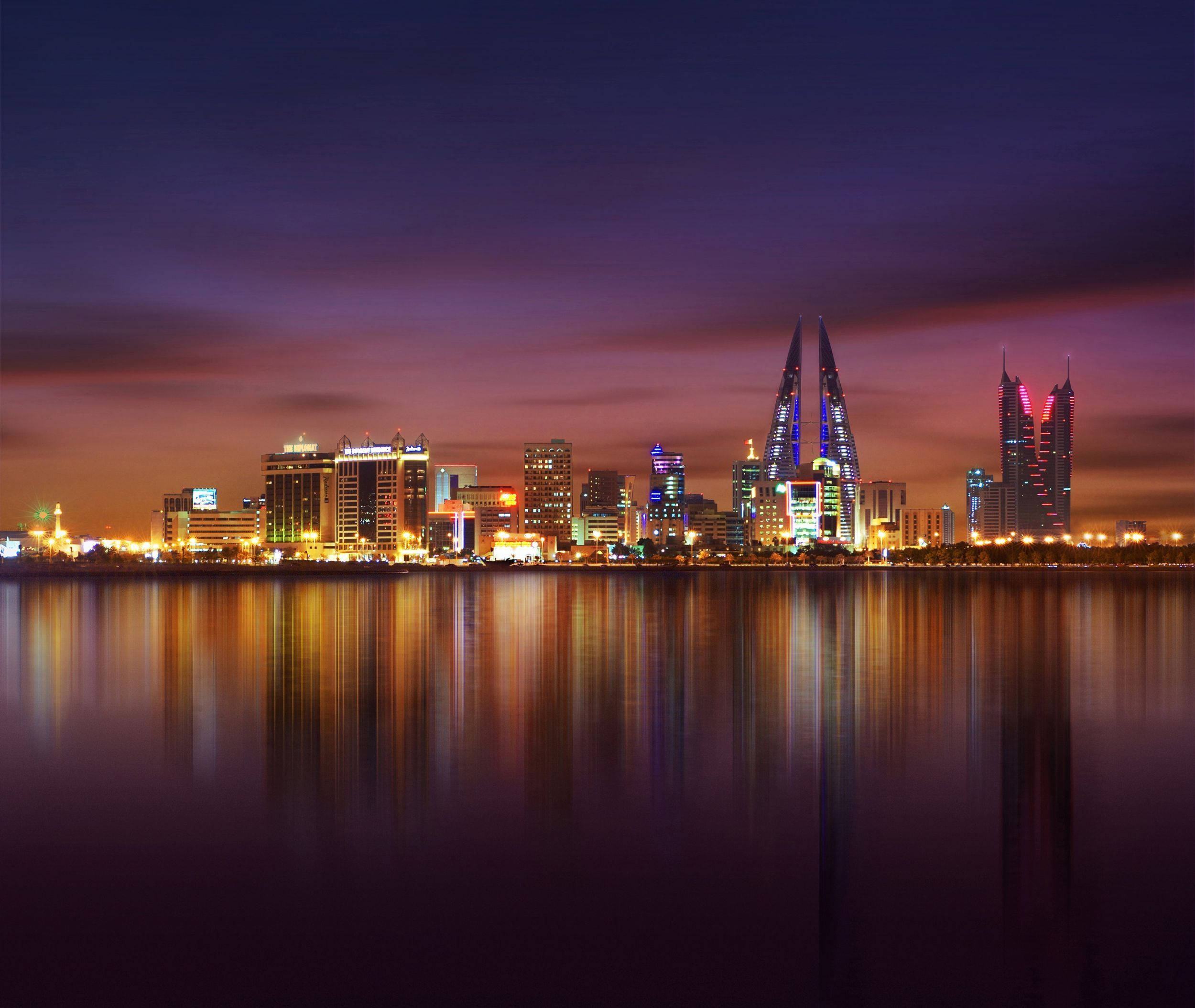 Bahrain 桌布 壁紙 — 黃昏時分充滿活力的城市天際線，色彩繽紛的摩天大樓在紫色天空下倒映在平靜的水面上。