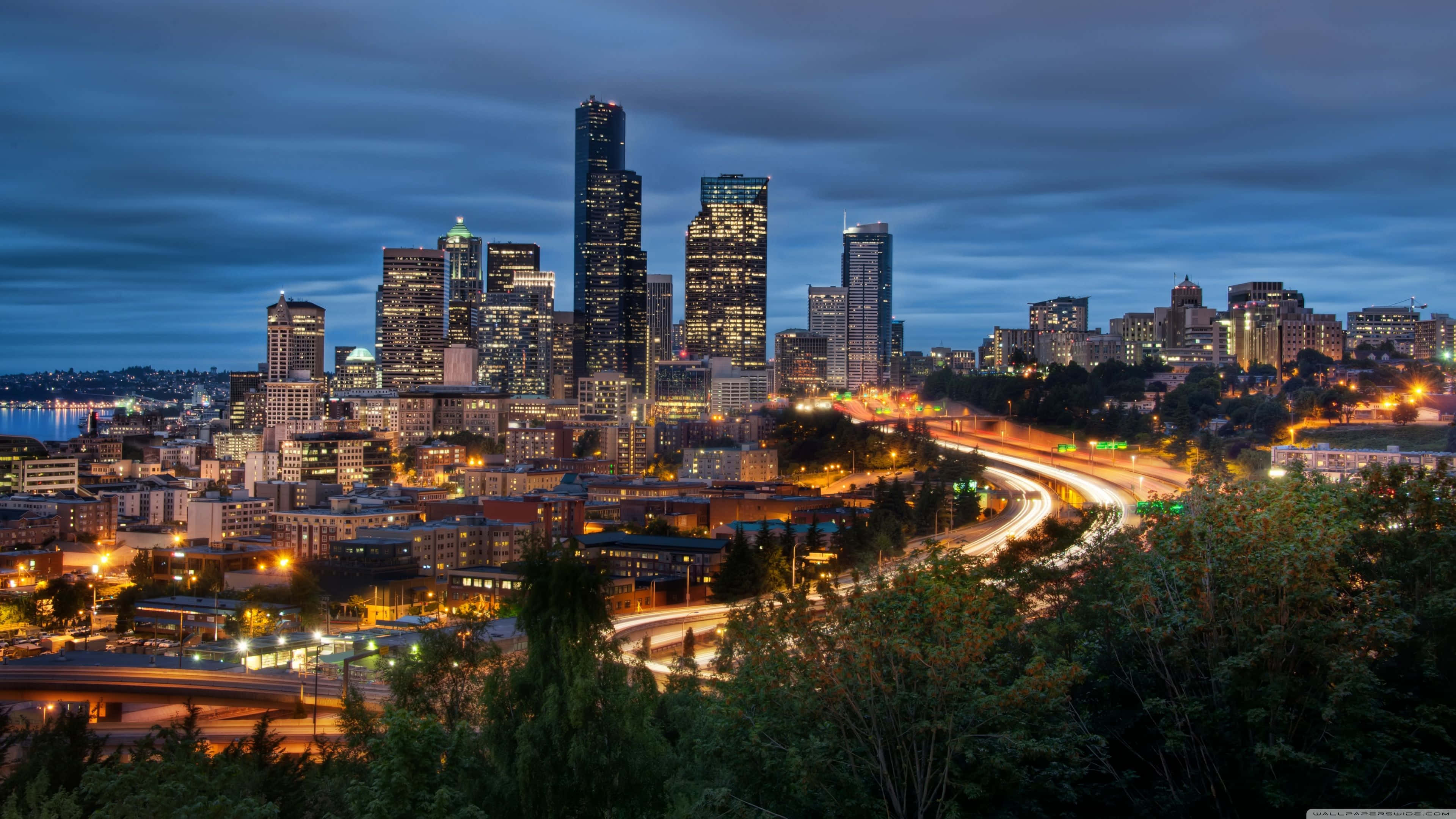 Seattle Washington 桌布 壁紙 — 黃昏時分充滿活力的城市天際線，閃爍的摩天大樓和蜿蜒穿過茂密綠化的交通光跡。