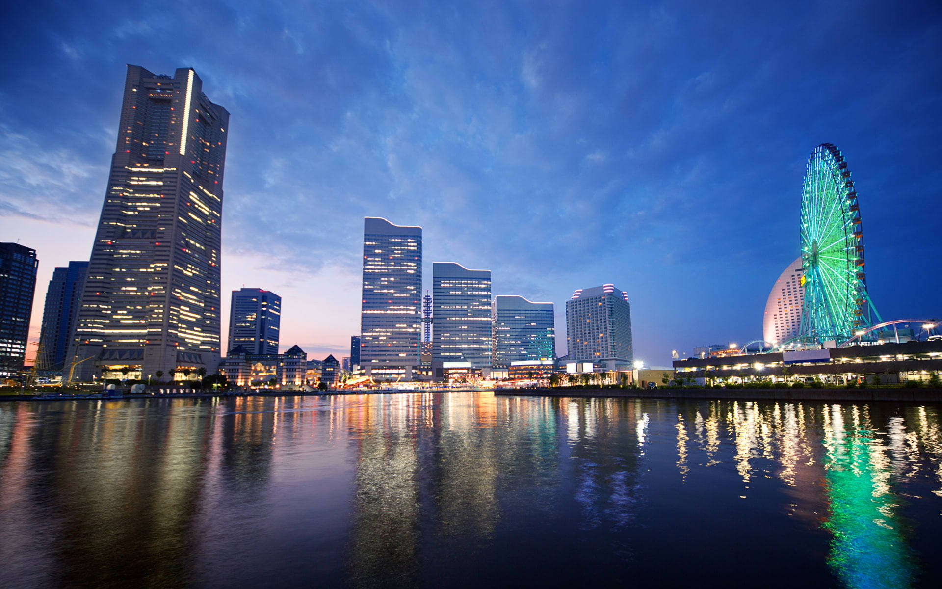 Yokohama 桌布 壁紙 — 黃昏時分充滿活力的城市天際線，閃爍的摩天大樓和色彩繽紛的摩天輪倒映在平靜的水面上。