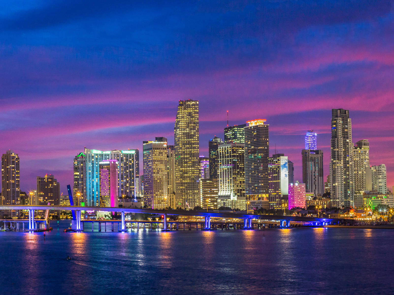 Miami 桌布 壁紙 — 黃昏時分充滿活力的城市天際線，閃爍的摩天大樓和色彩繽紛的天空倒映在平靜的水面上。