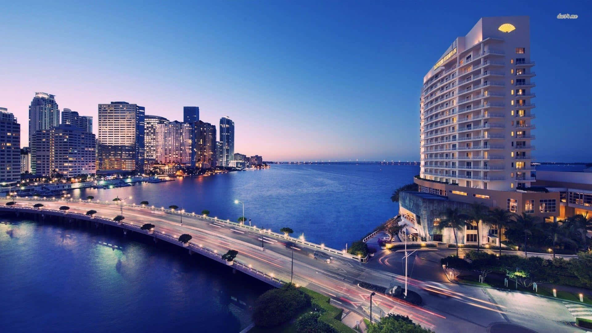 Miami 桌布 壁紙 — 黃昏時分充滿活力的城市天際線，閃爍的摩天大樓，帶有光跡的海濱道路，以及平靜水域旁的醒目酒店。