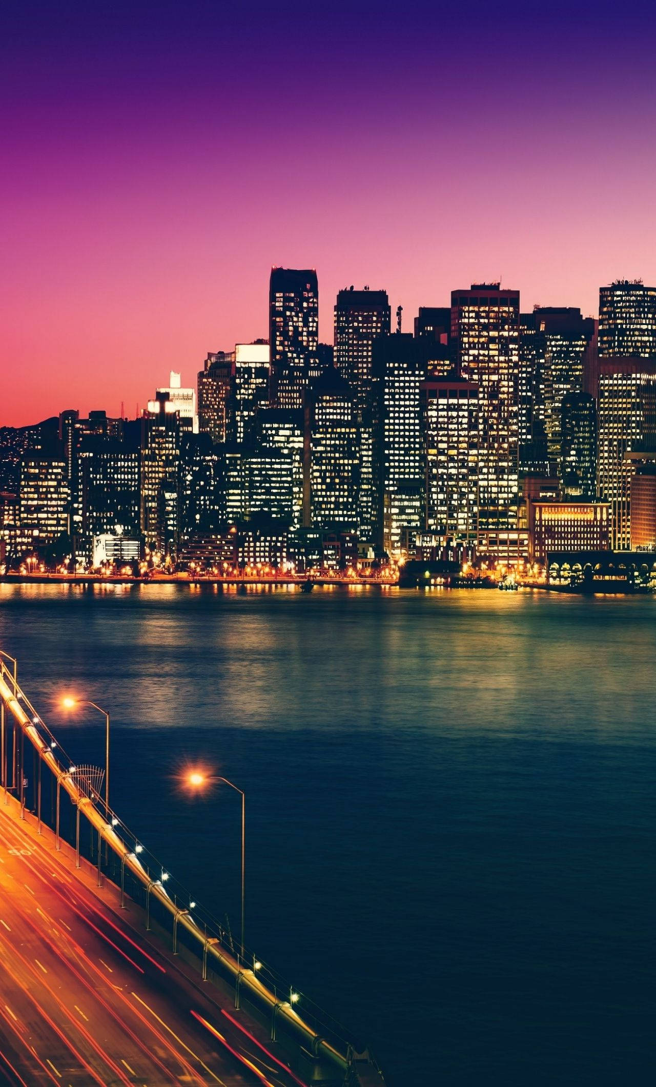 san francisco iPhone 桌布 壁紙 — 黃昏時分充滿活力的城市天際線，閃爍的摩天大樓倒映在平靜的水面上，下方橋樑的光線條紋。