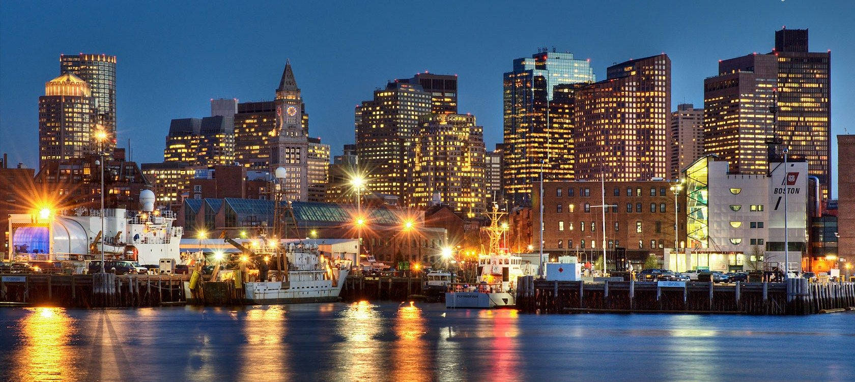 Boston 桌布 壁紙 — 黃昏時分充滿活力的城市天際線，閃爍的摩天大樓和停泊的船隻倒映在平靜的水面上。