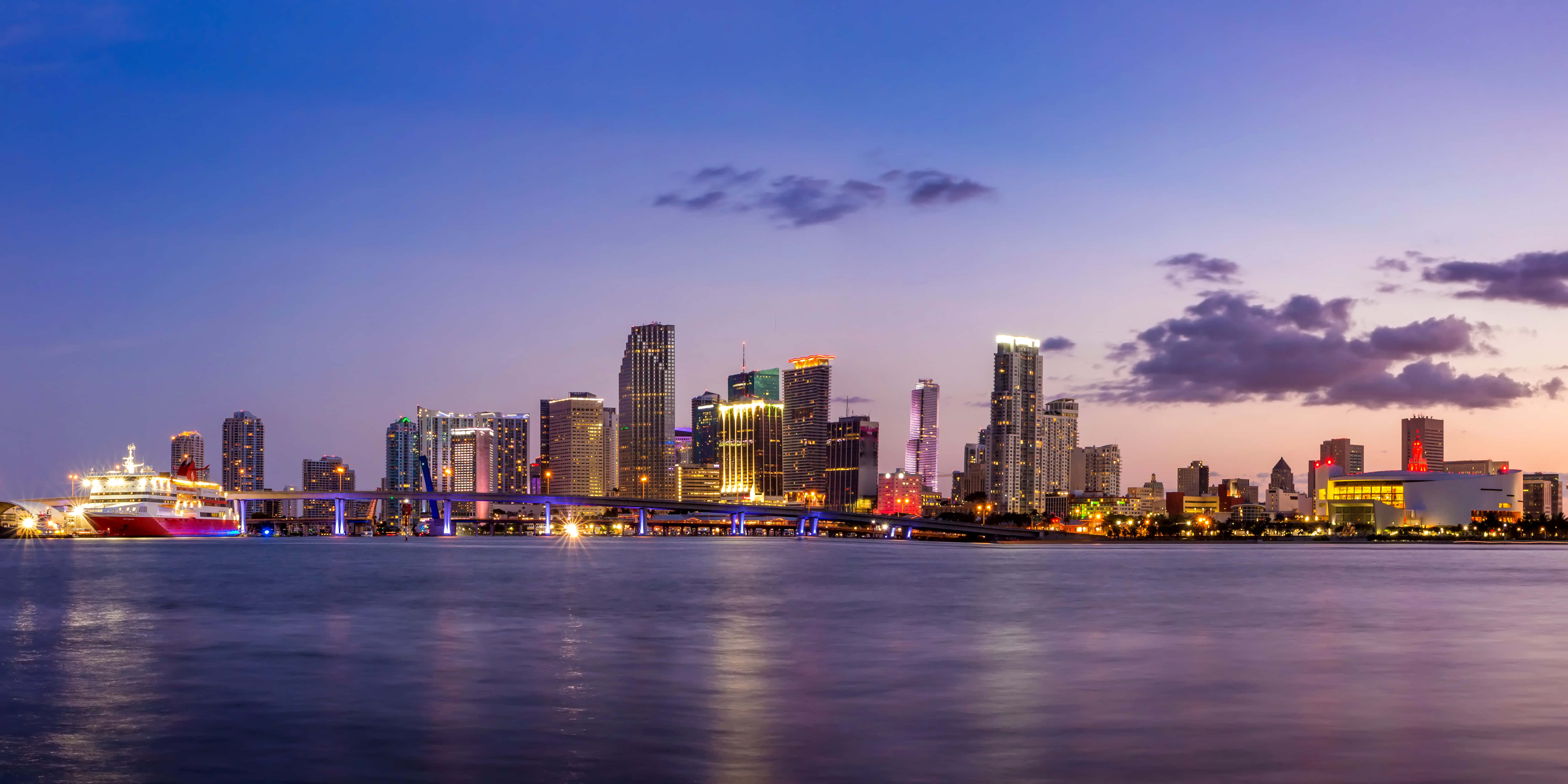 Miami 4K 桌布 壁紙 — 黃昏時分充滿活力的城市天際線，閃爍的摩天大樓和遊輪在紫色雲層下平靜的水面上倒映。