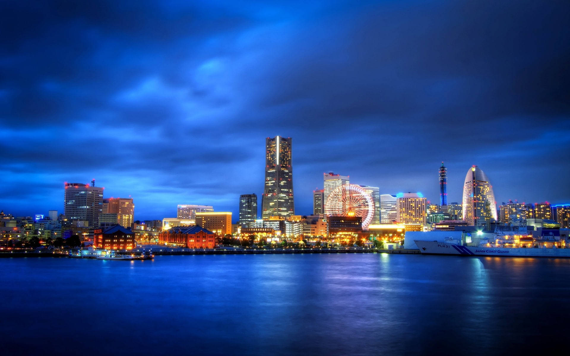 Yokohama 桌布 壁紙 — 黃昏時分充滿活力的城市天際線，閃爍的摩天大樓和摩天輪倒映在平靜的水面上，天空深藍多雲。