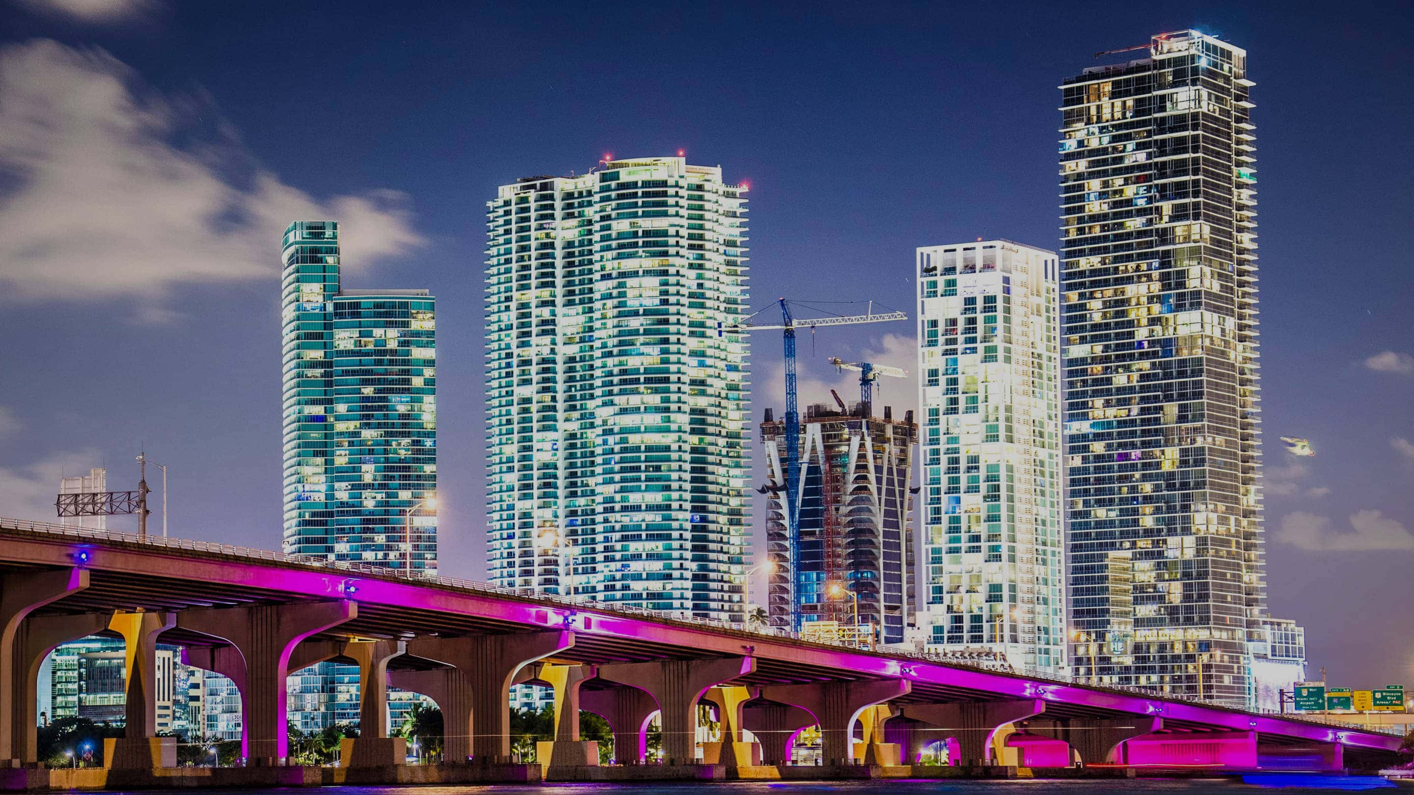 Miami 桌布 壁紙 — 黃昏時分充滿活力的城市天際線，閃爍的摩天大樓和一座橫跨水道的洋紅色橋樑。