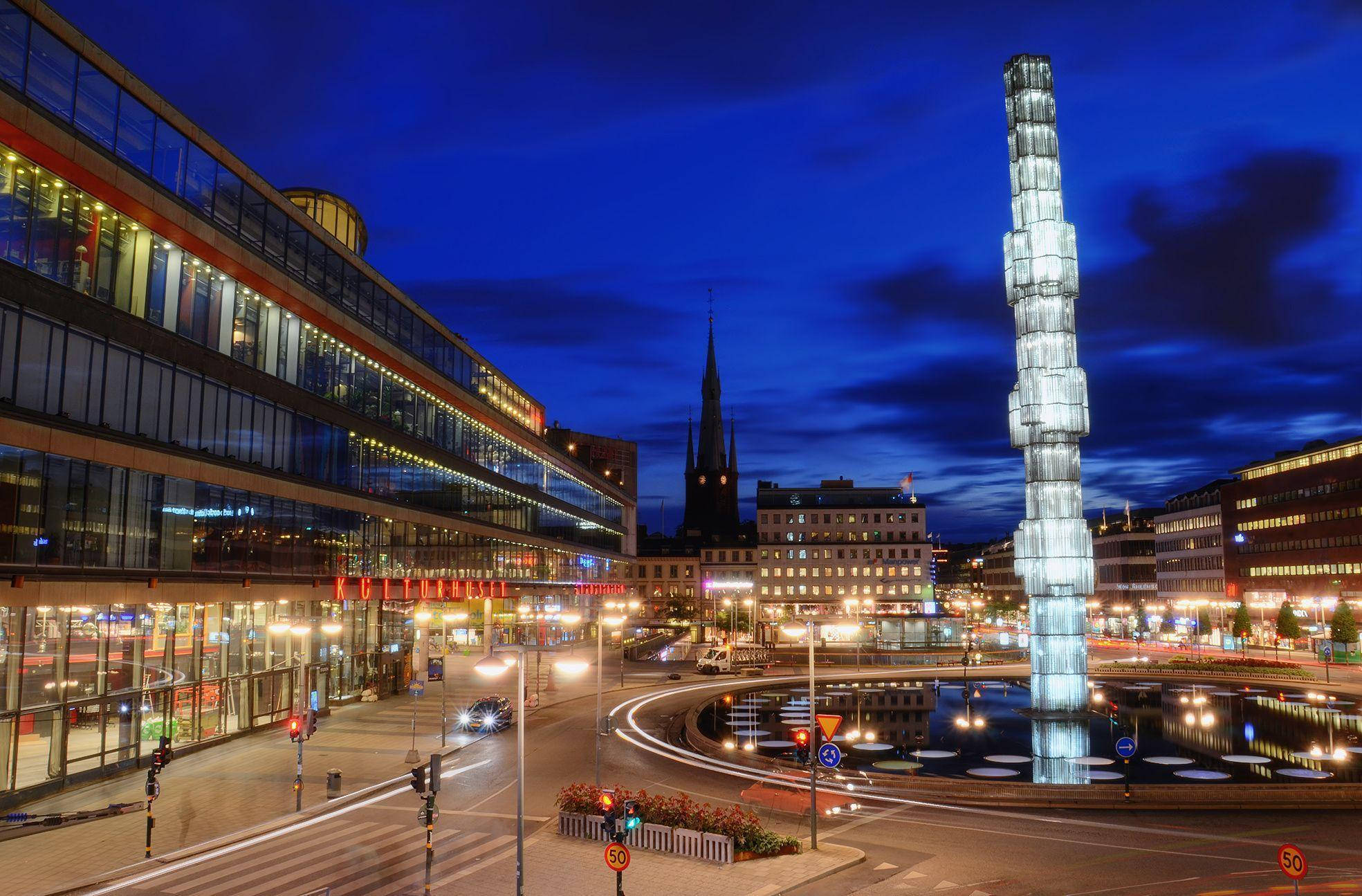 Sweden 桌布 壁紙 — 黃昏時分充滿活力的城市景觀，一座發光的現代塔樓倒映在圓形噴泉中，周圍是燈火通明的建築物和交通燈跡。