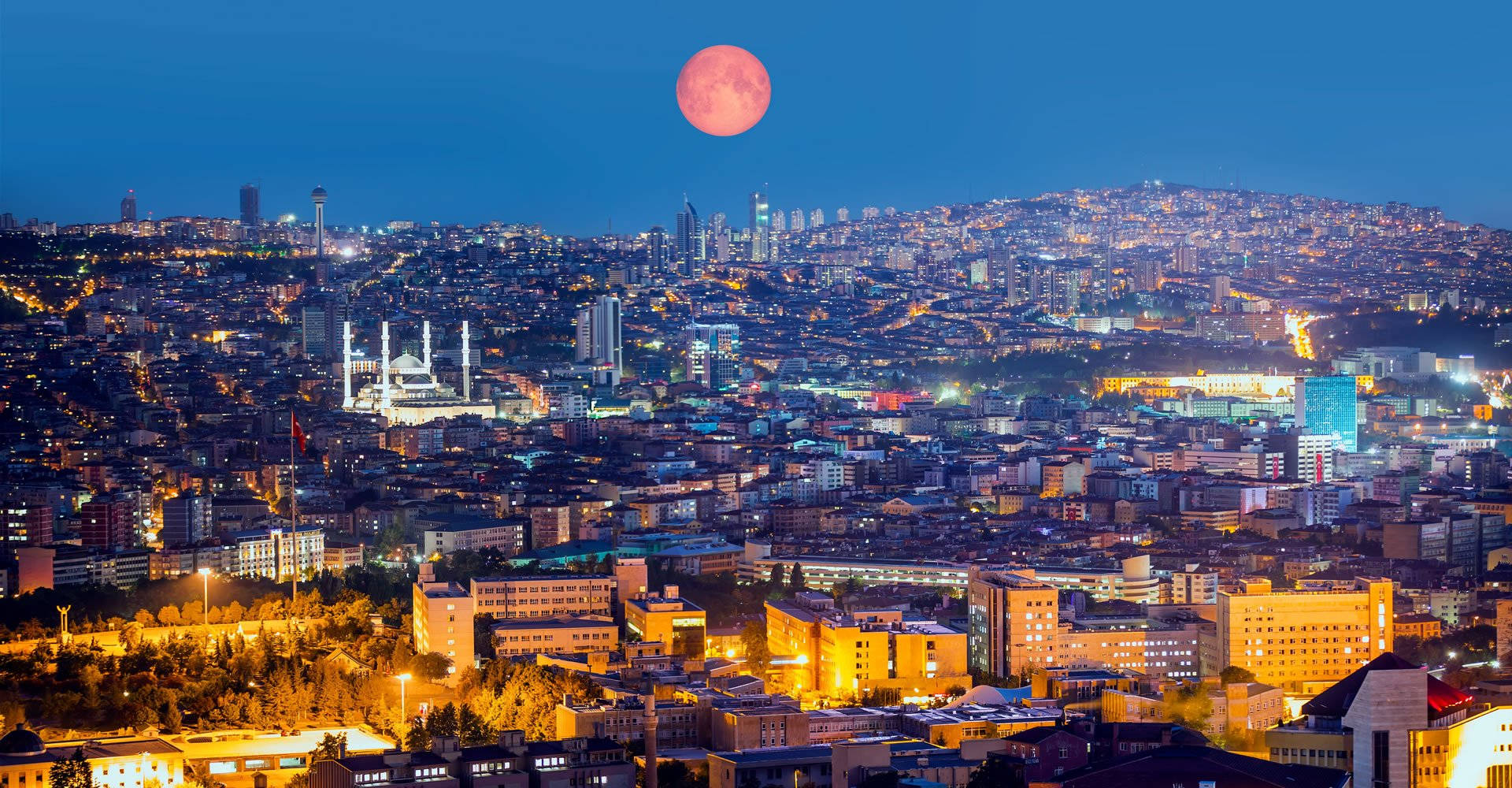 Ankara 桌布 壁紙 — 黃昏時分充滿活力的城市景觀，一輪發光的紅月懸掛在燈火通明的建築物和蜿蜒的街道之上。