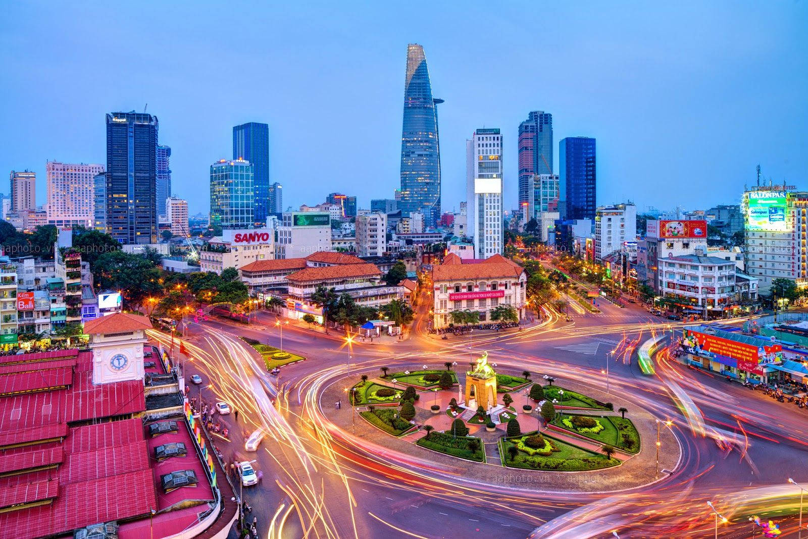Ho Chi Minh City 桌布 壁紙 — 黃昏時分充滿活力的城市景觀，交通燈光軌跡圍繞著一個被現代摩天大樓和歷史建築環繞的中央圓環旋轉。