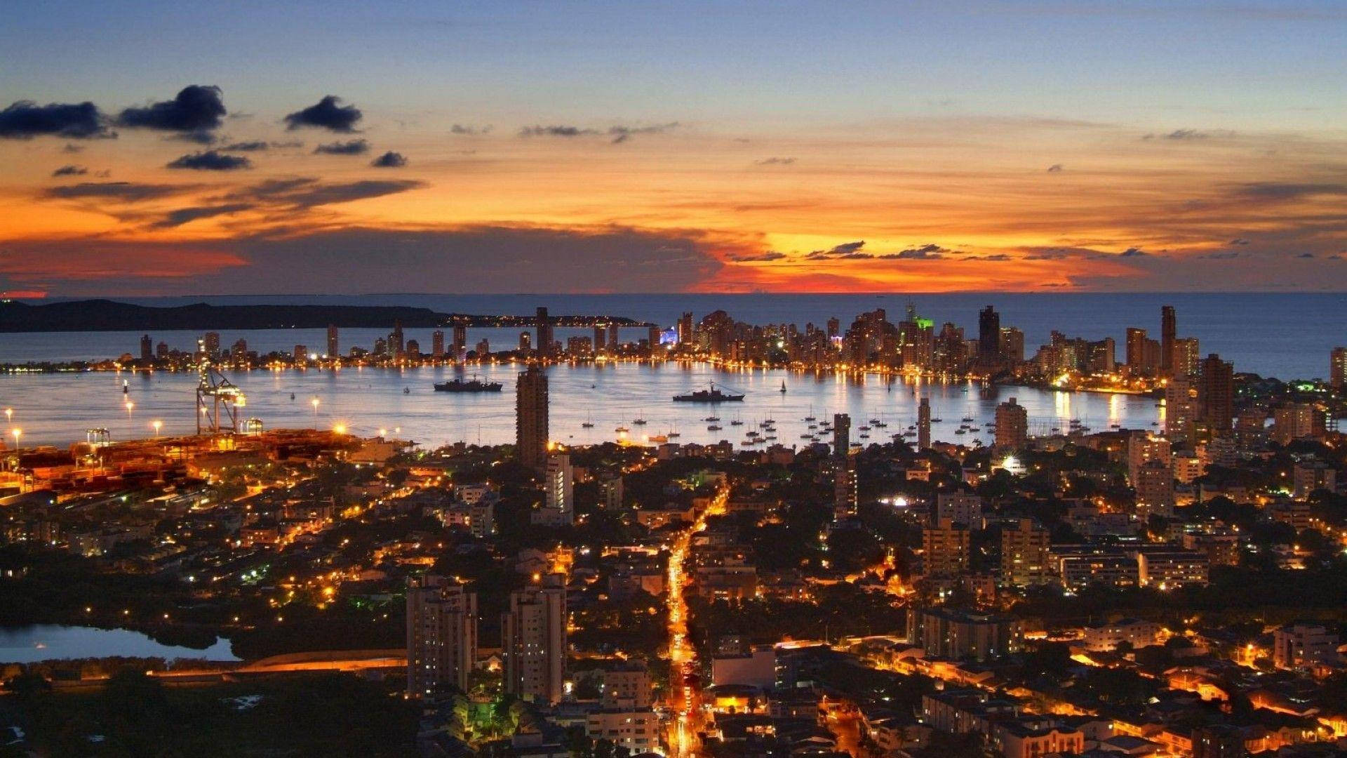 Colombia 桌布 壁紙 — 黃昏時分充滿活力的城市景觀，在火紅橙和紫色天空下，閃爍的摩天大樓和水面上的船隻。