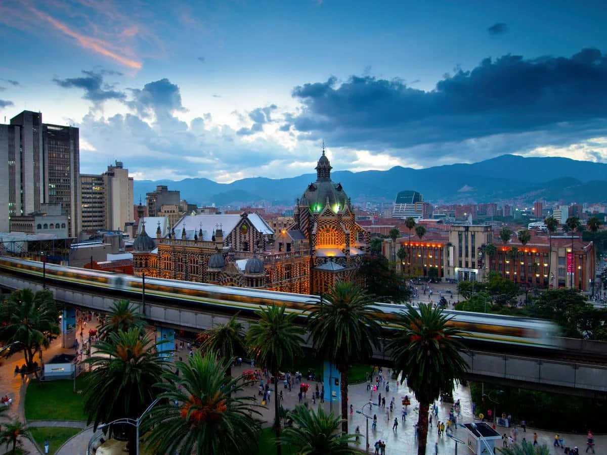 Colombia 桌布 壁紙 — 黃昏時分充滿活力的城市景觀，有一座發光的歷史建築、棕櫚樹和一列在戲劇性多雲天空下行駛的火車。