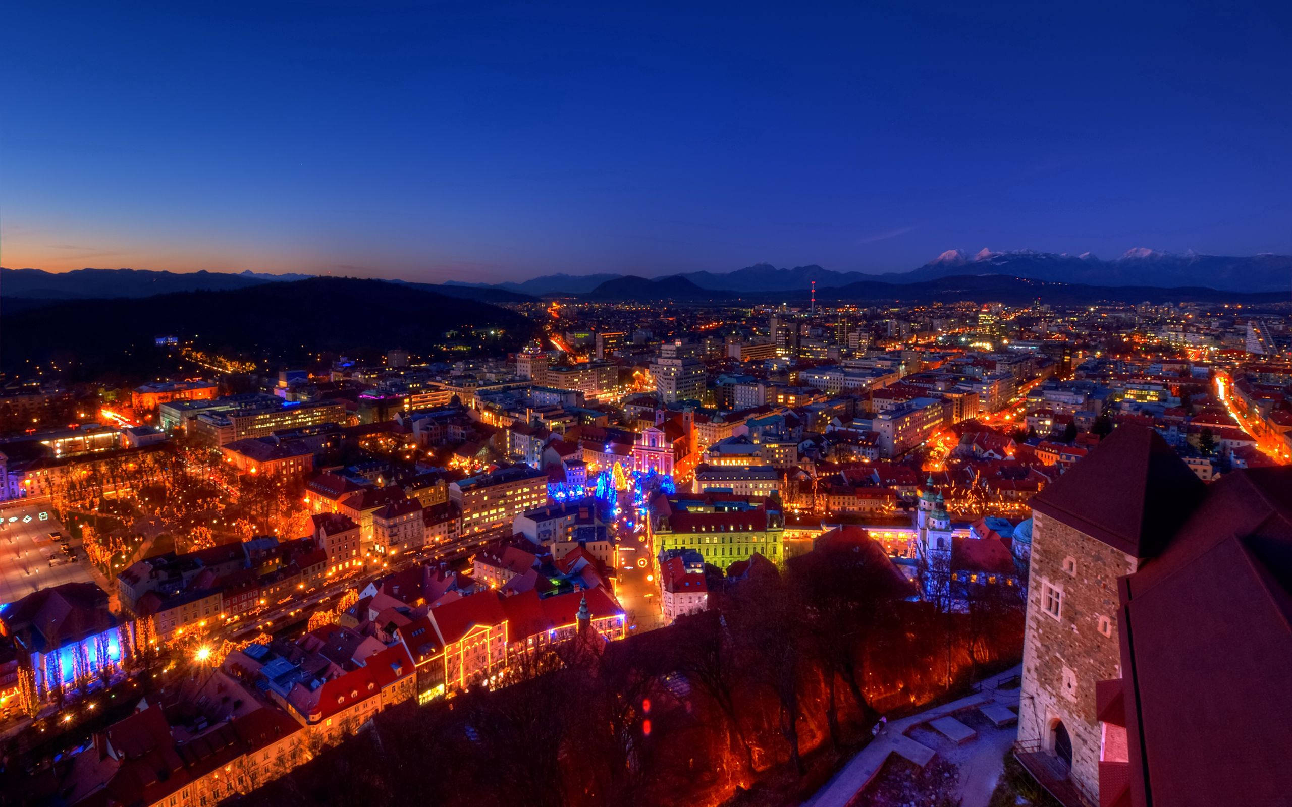 Slovenia 桌布 壁紙 — 黃昏時分充滿活力的城市景觀，橙色和藍色的燈光照亮歷史建築和蜿蜒的街道，映襯著深藍色的天空。