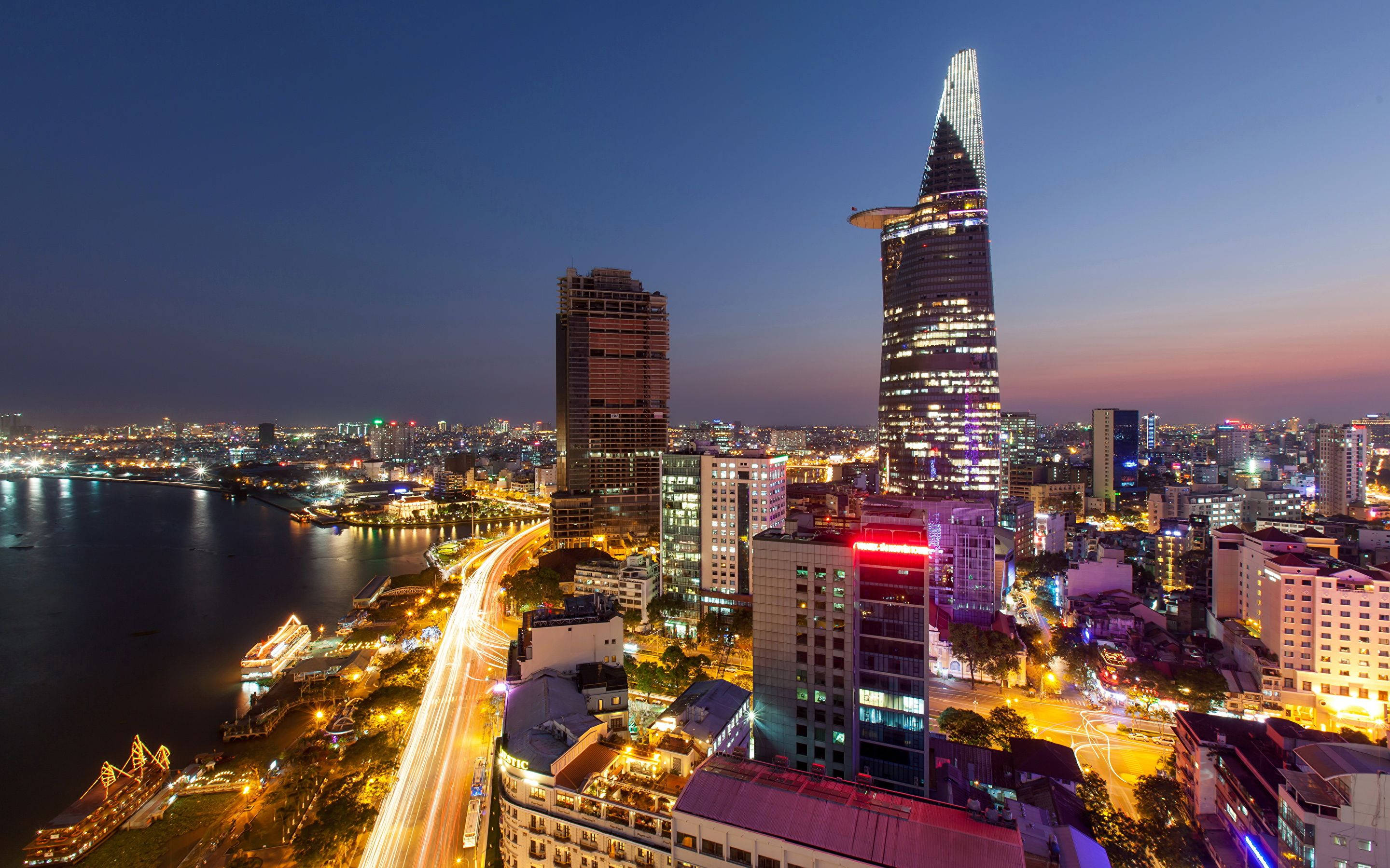 Ho Chi Minh City 桌布 壁紙 — 黃昏時分充滿活力的城市景觀，海濱沿岸有燈火通明的摩天大樓和發光的車流痕跡。
