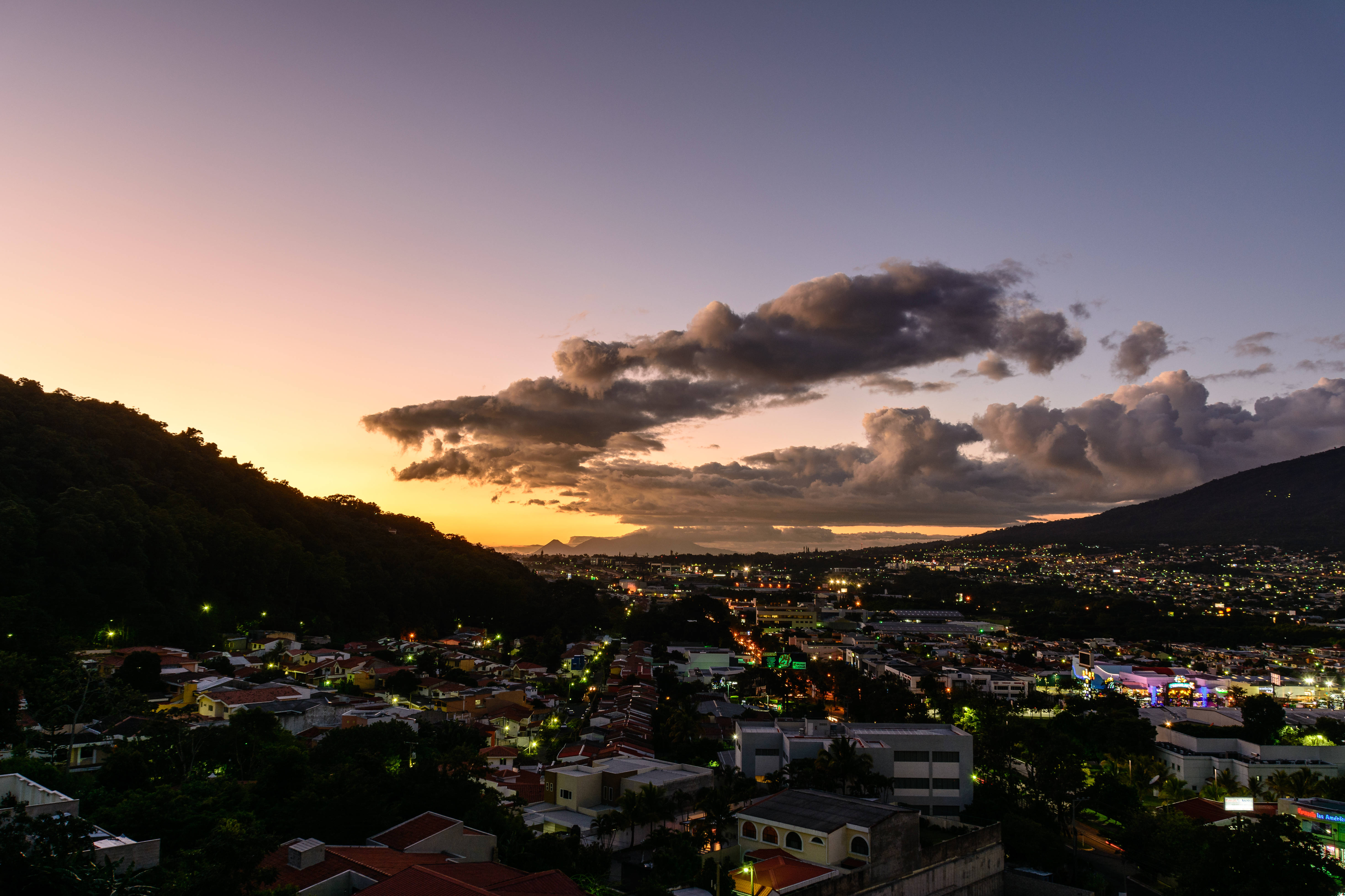El Salvador 桌布 壁紙 — 黃昏時分充滿活力的城市景觀，溫暖的橙色夕陽色調照亮了廣闊城市山谷上方的雲層，山谷中點綴著閃爍的路燈。