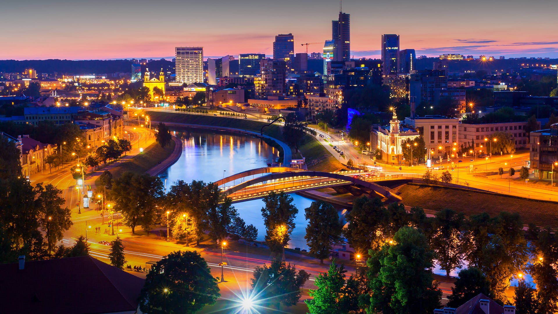 Vilnius 桌布 壁紙 — 黃昏時分充滿活力的城市景觀，發光的河流蜿蜒穿過燈火通明的摩天大樓和橋樑。