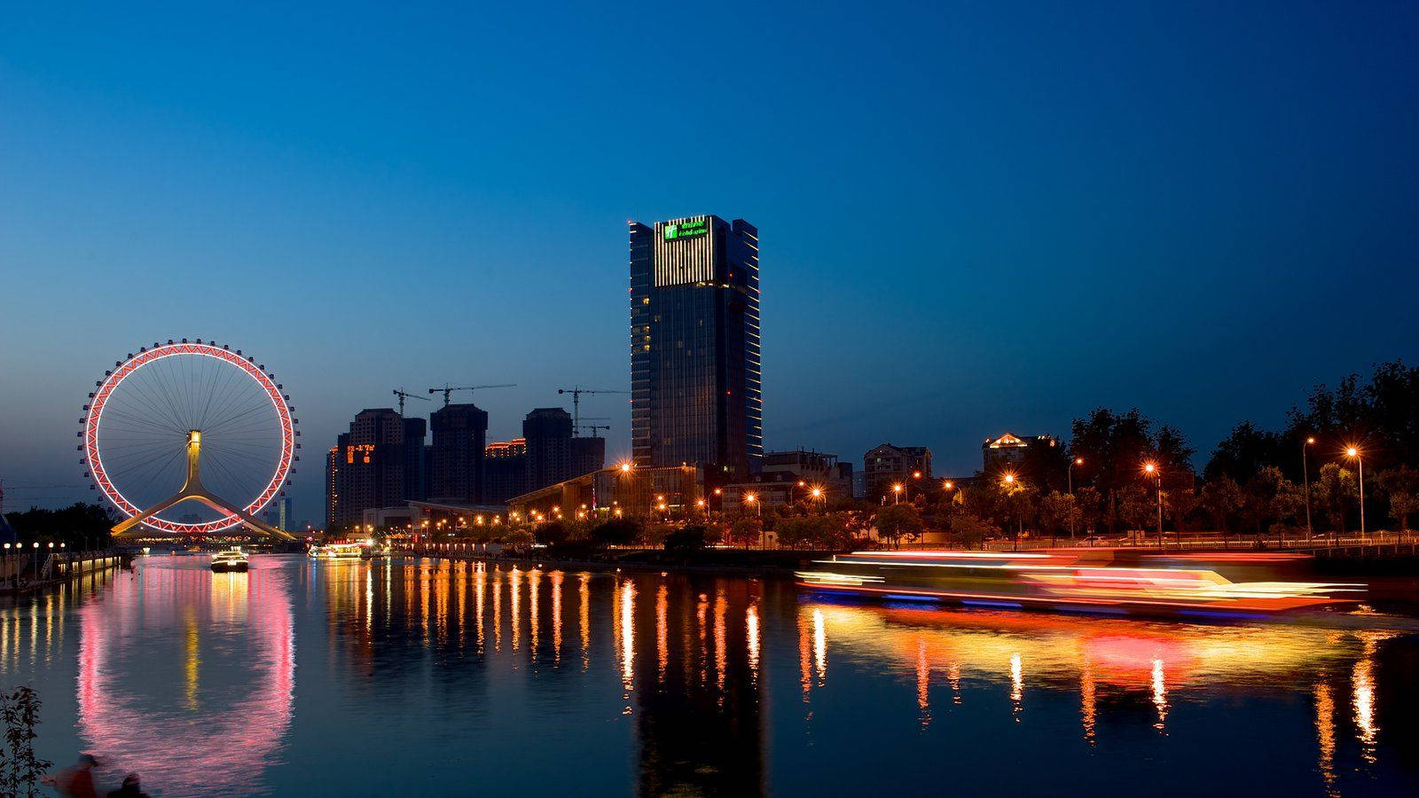 Tianjin 桌布 壁紙 — 黃昏時分充滿活力的城市景觀，發光的摩天輪和摩天大樓倒映在河中，船隻帶著光跡疾馳而過。