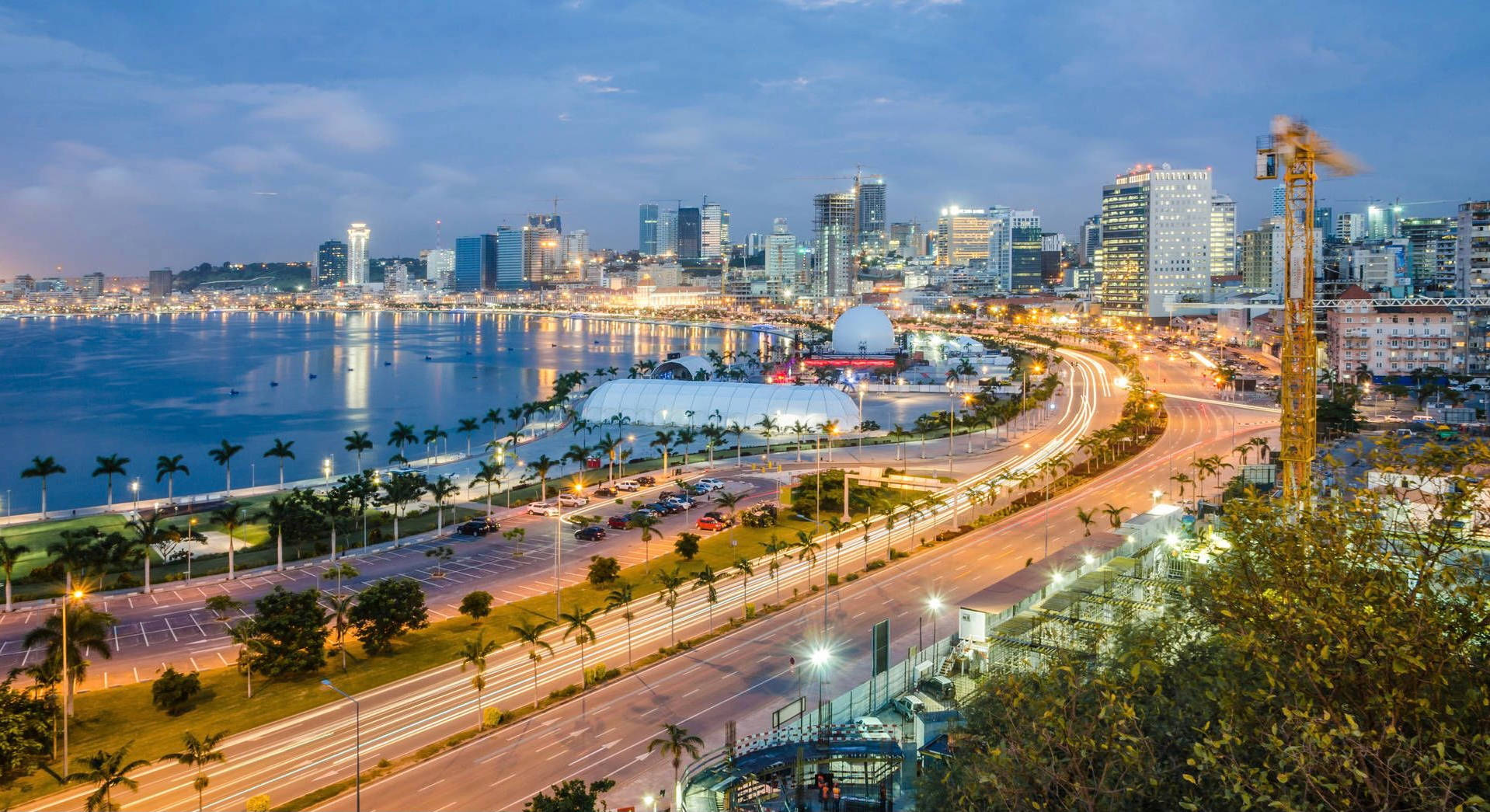 Angola 桌布 壁紙 — 黃昏時分充滿活力的城市景觀，閃爍的摩天大樓、棕櫚樹成蔭的道路以及沿海濱的車流光跡。