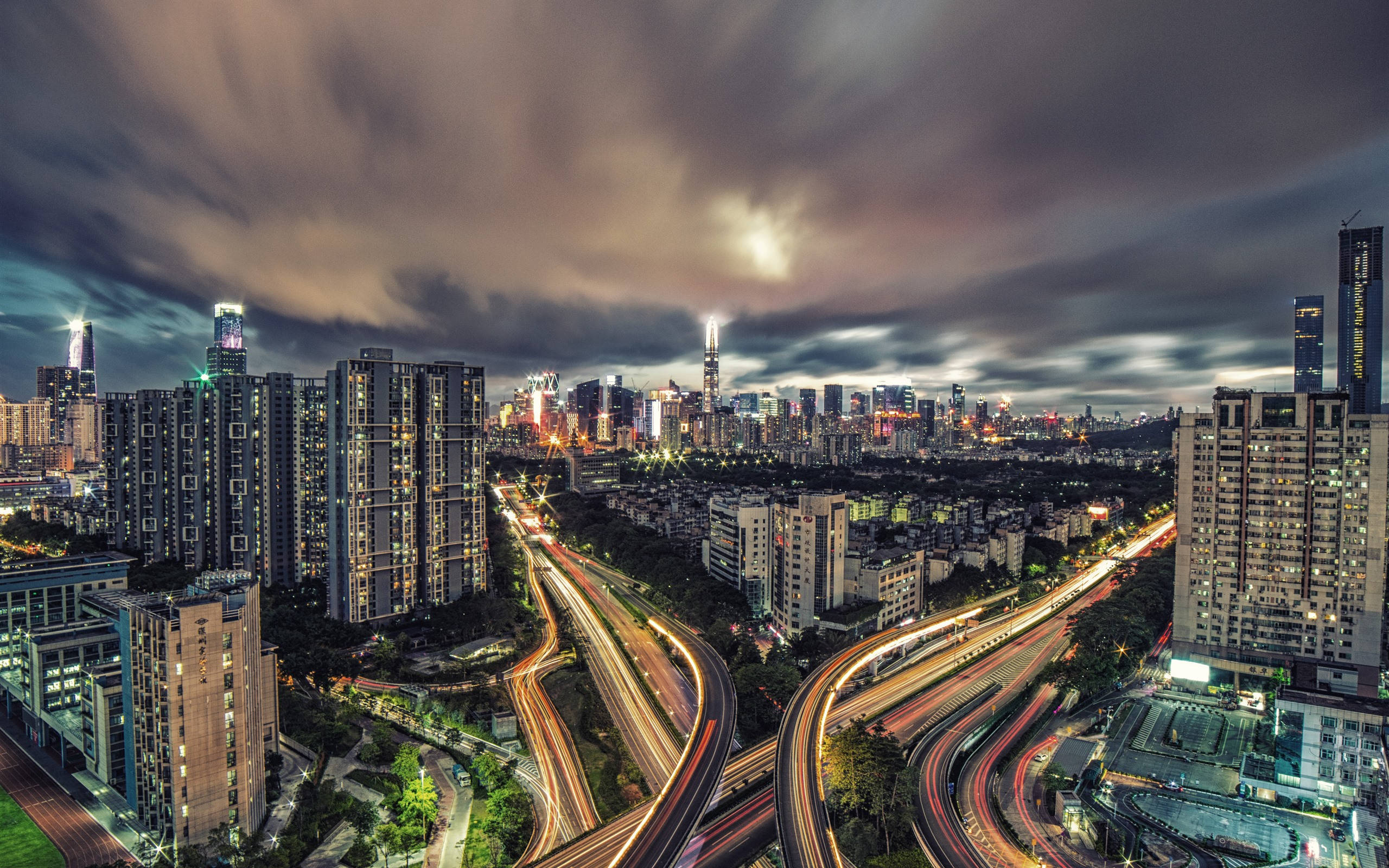 Shenzhen 桌布 壁紙 — 黃昏時分充滿活力的城市景觀，閃爍的摩天大樓和車流的光跡在戲劇性的雲層下。