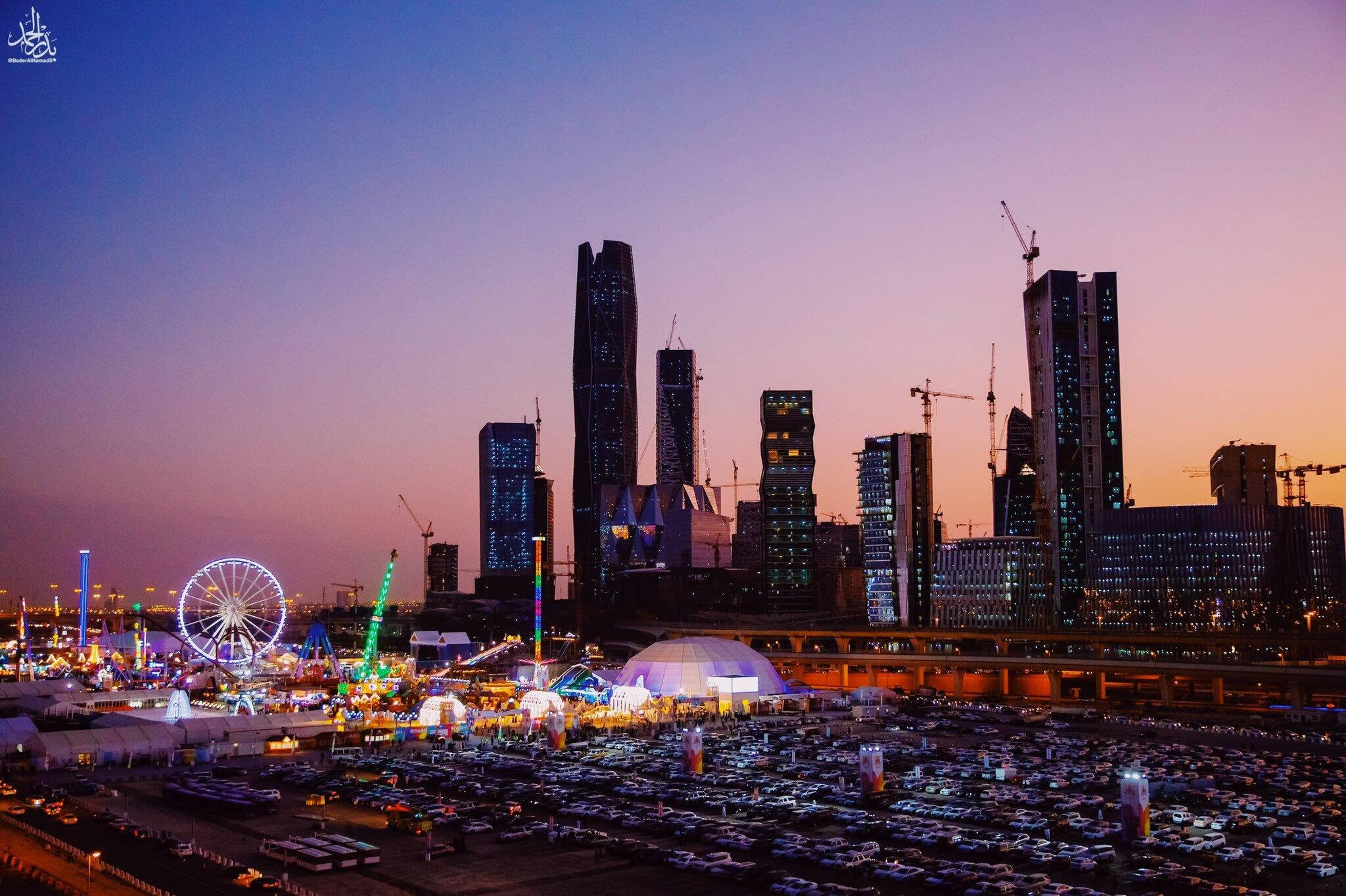 Riyadh 桌布 壁紙 — 黃昏時分充滿活力的城市景觀，閃爍的摩天大樓和色彩繽紛的遊樂園，設有摩天輪。