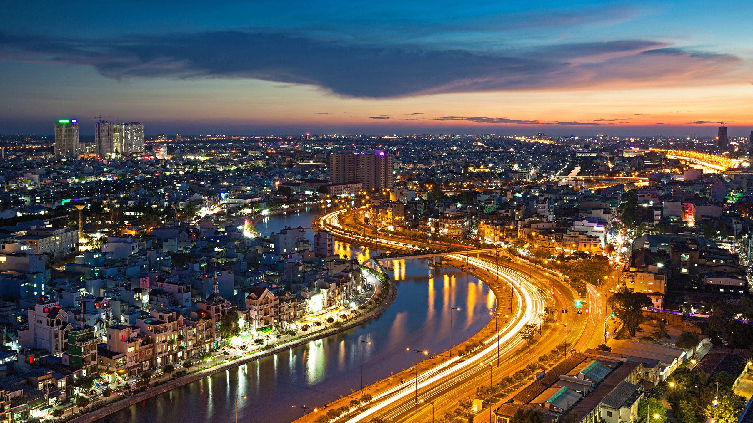 Ho Chi Minh City 桌布 壁紙 — 黃昏時分充滿活力的城市景觀，閃爍的燈光倒映在蜿蜒的河流和繁忙的高速公路上。