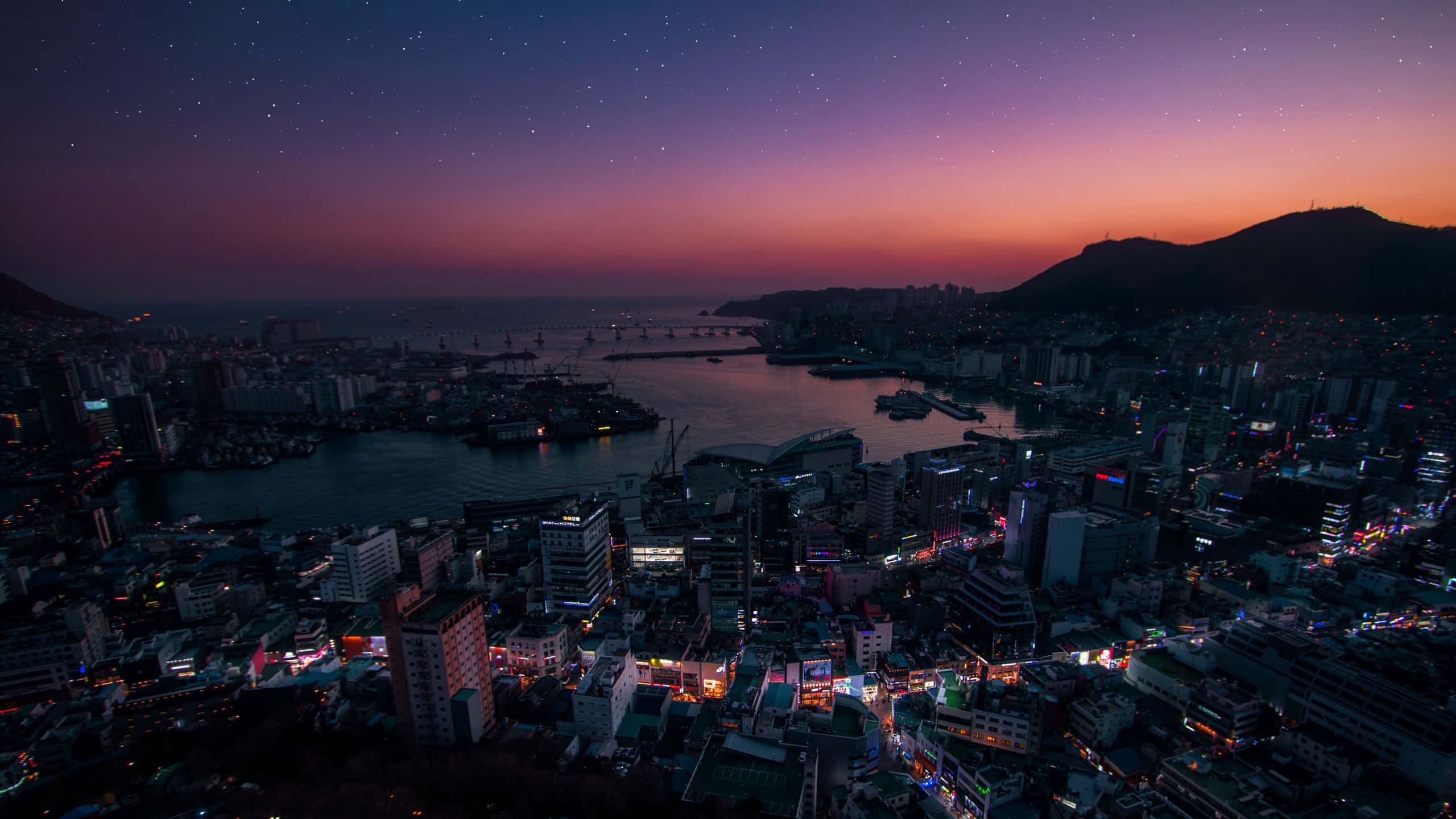 南韓 桌布 壁紙 — 黃昏時分充滿活力的城市景觀，霓虹燈在紫色和橙色星空下的水面上閃爍倒映。