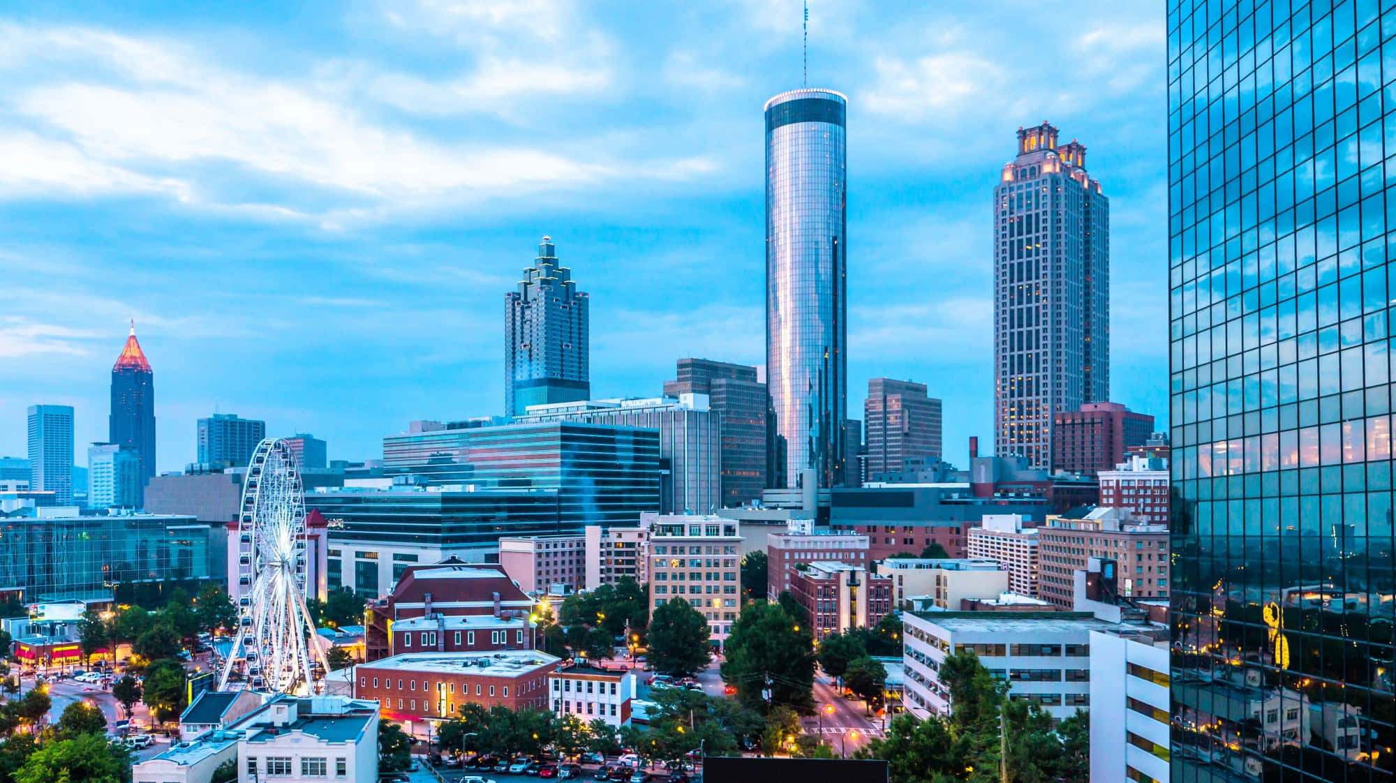 Atlanta Georgia 桌布 壁紙 — 黃昏時分充滿活力的城市景觀，高聳的摩天大樓、閃爍的摩天輪和溫暖的路燈映襯著柔和的藍天。
