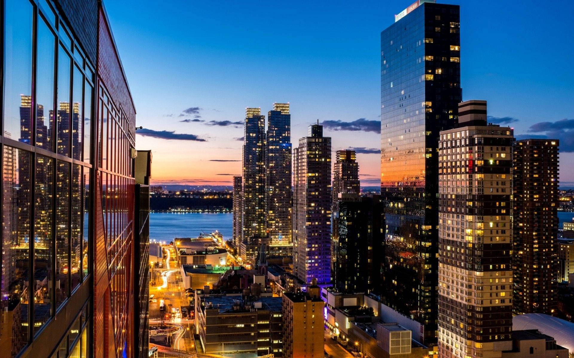 Nyc Aesthetic 桌布 壁紙 — 黃昏時分充滿活力的城市景觀，高聳的摩天大樓在閃爍的海濱上空反射出橙色和藍色的天空。