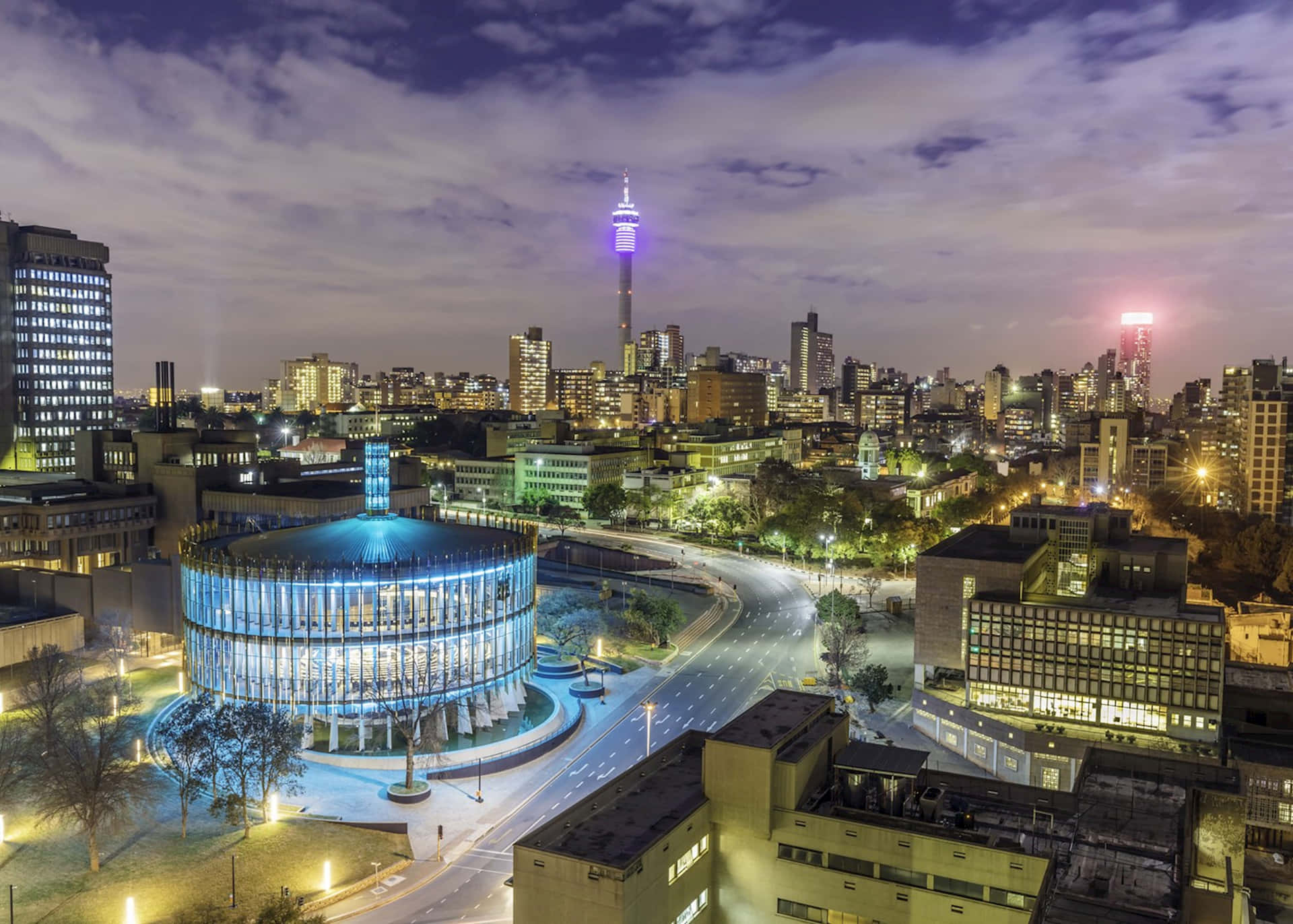 South Africa 桌布 壁紙 — 黃昏時分充滿活力的城市景觀鳥瞰圖，現代建築燈火通明，發光的塔樓映襯著紫色的天空。