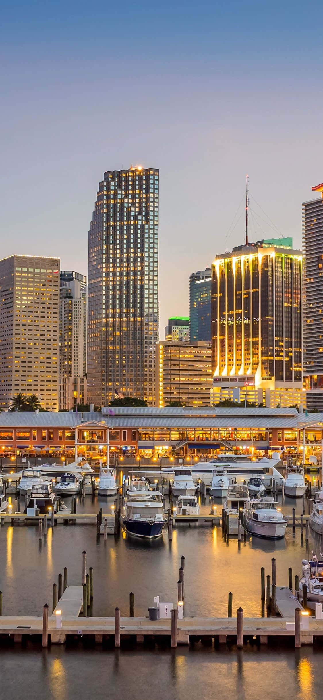Miami iPhone 桌布 壁紙 — 黃昏時分充滿活力的城市港口，閃爍的摩天大樓倒映在平靜的水面上，周圍停泊著船隻。