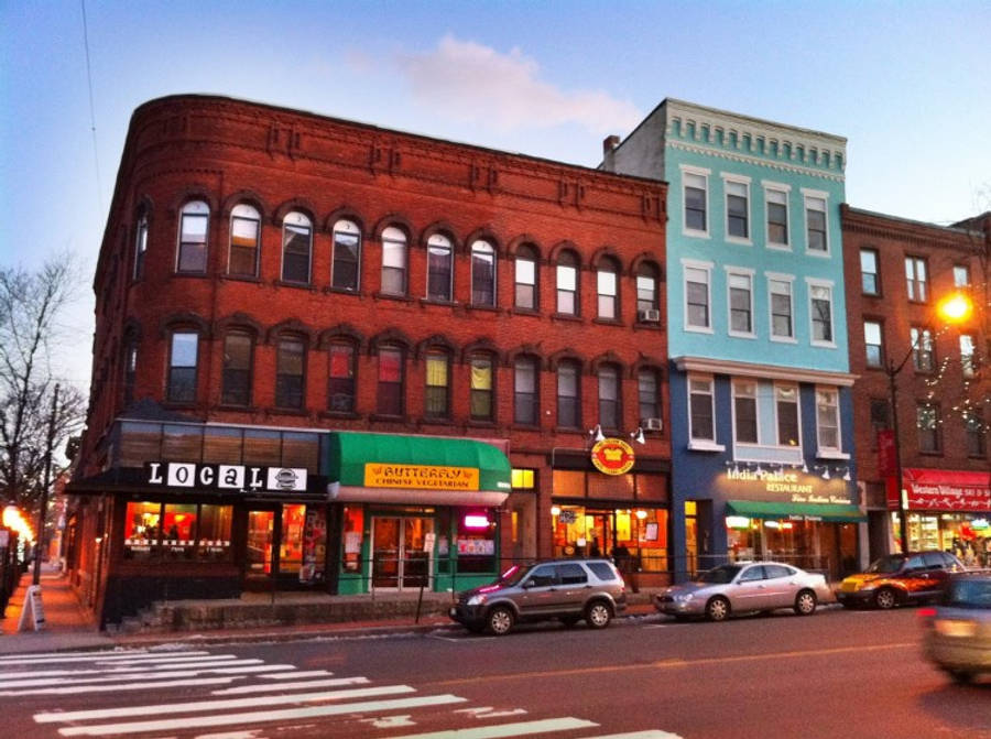 Massachusetts 桌布 壁紙 — 黃昏時分充滿活力的城市街道，色彩繽紛的磚砌建築、發光的店面以及停在柔和藍天下的汽車。
