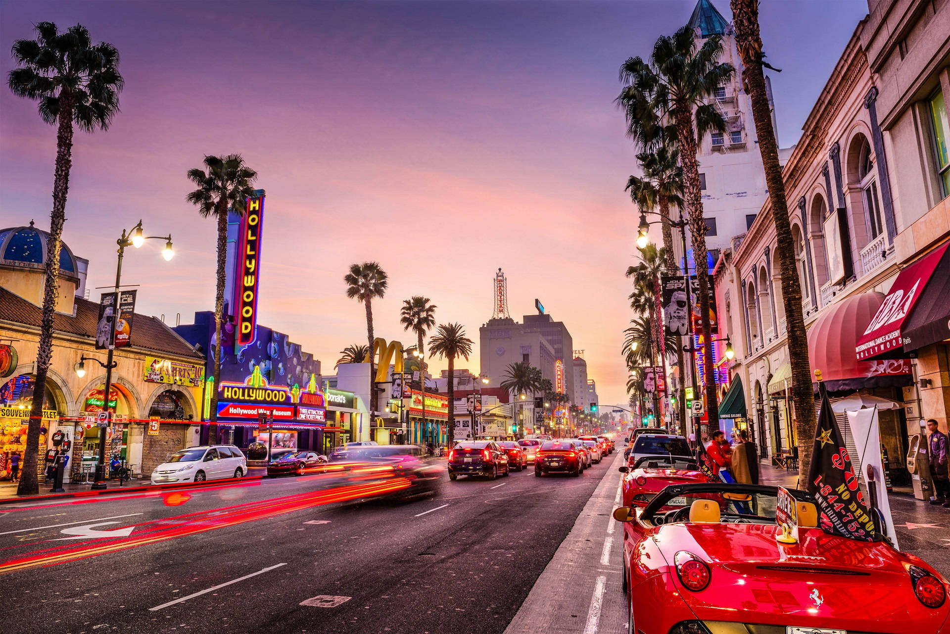 hollywood street 桌布 壁紙 — 黃昏時分充滿活力的好萊塢大道，棕櫚樹、霓虹燈招牌和汽車燈光在粉紫色天空下劃過。