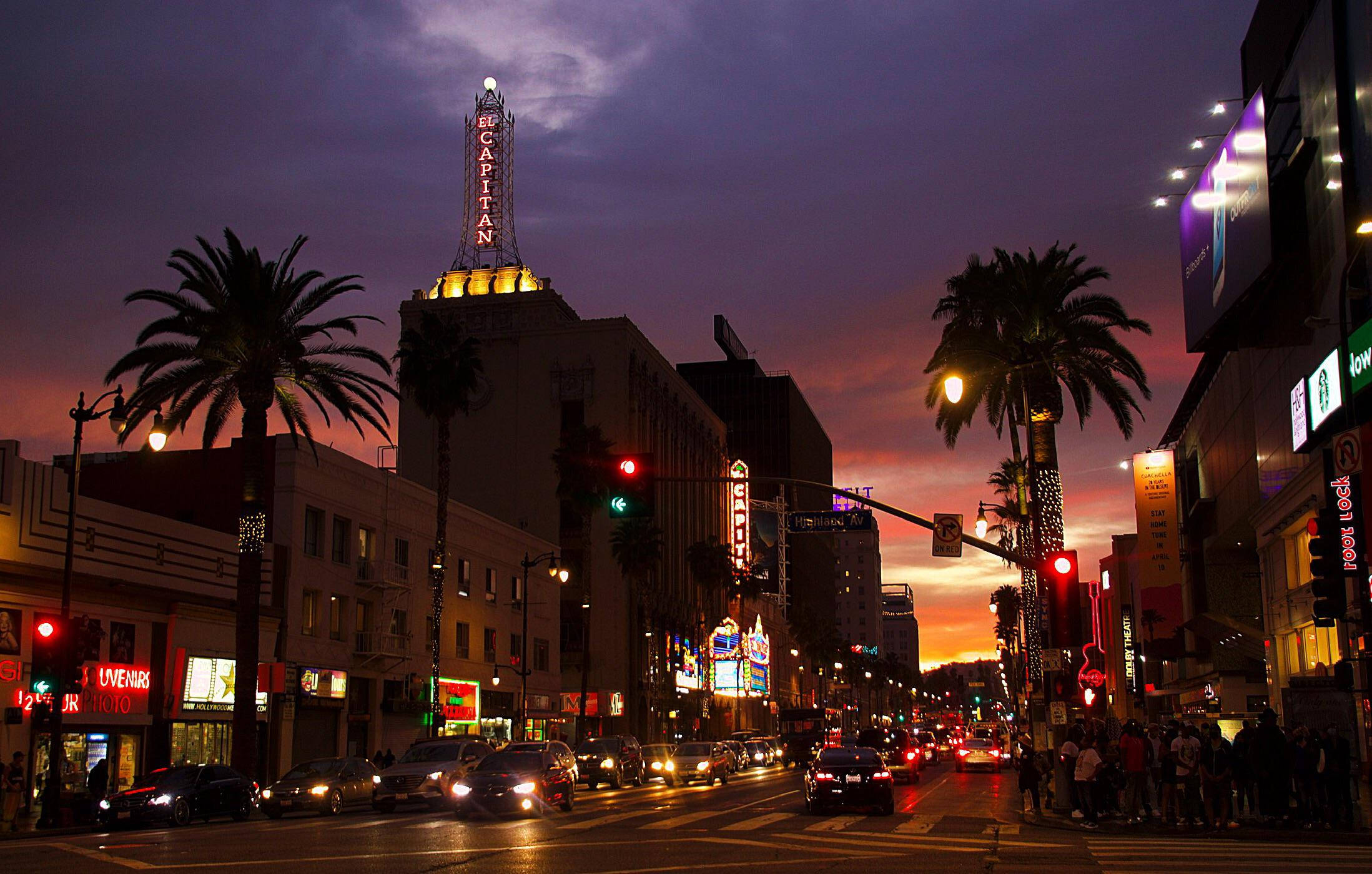 hollywood street 桌布 壁紙 — 黃昏時分充滿活力的好萊塢街道，棕櫚樹、發光的霓虹燈招牌，以及在紫橙色天空下流動的交通。