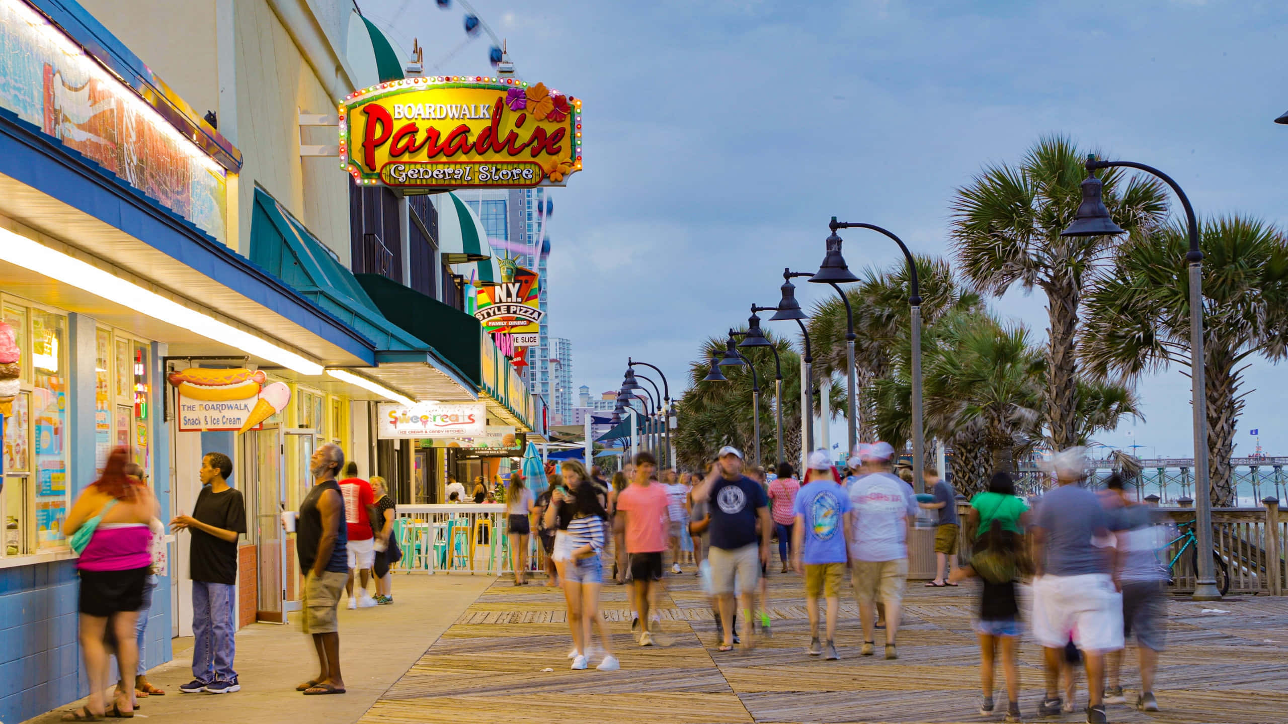 Myrtle Beach 桌布 壁紙 — 黃昏時分充滿活力的木板路，棕櫚樹、發光的霓虹燈招牌，以及漫步經過糖果店和美食攤位的人群。