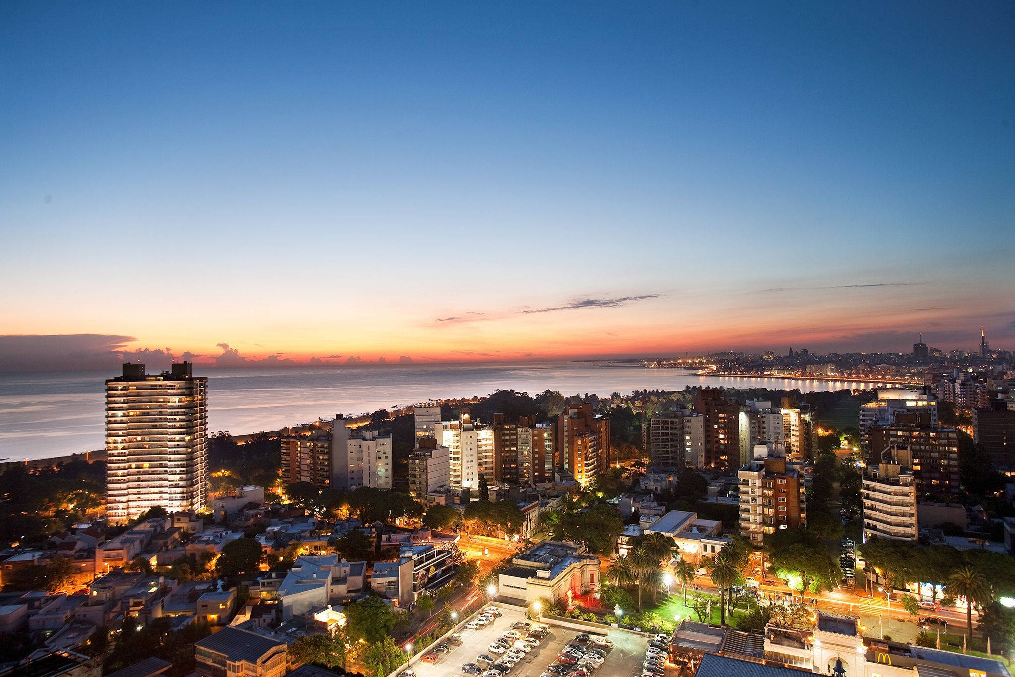Uruguay 桌布 壁紙 — 黃昏時分充滿活力的沿海城市景觀，溫暖的燈光從建築物中閃耀，映襯著海洋上方的漸變天空。