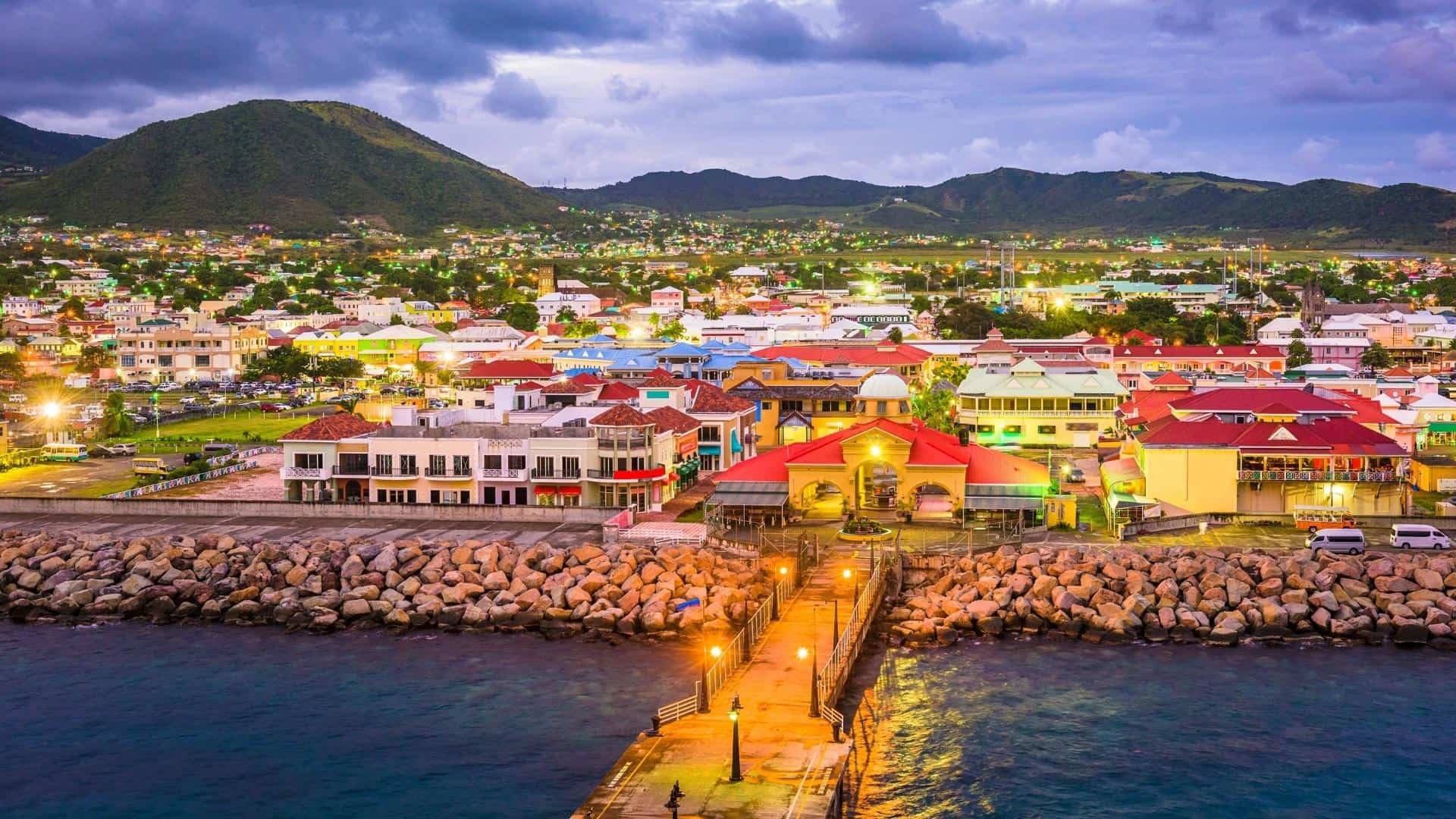 Caribbean 桌布 壁紙 — 黃昏時分充滿活力的沿海小鎮，色彩繽紛的建築物、石頭防波堤，以及延伸至平靜藍色海水的燈光碼頭。