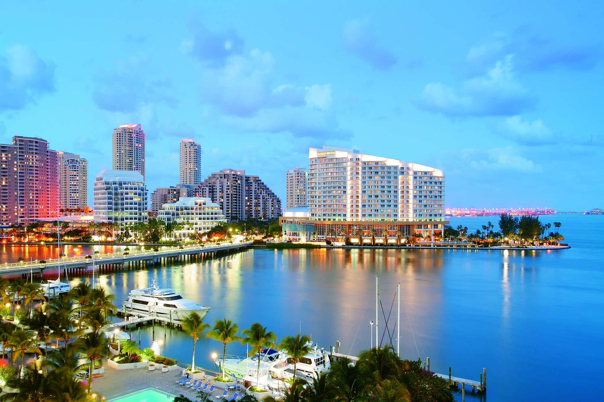 Miami 桌布 壁紙 — 黃昏時分充滿活力的海濱城市景觀，閃爍的摩天大樓倒映在平靜的水面上，周圍環繞著棕櫚樹和停泊的遊艇。
