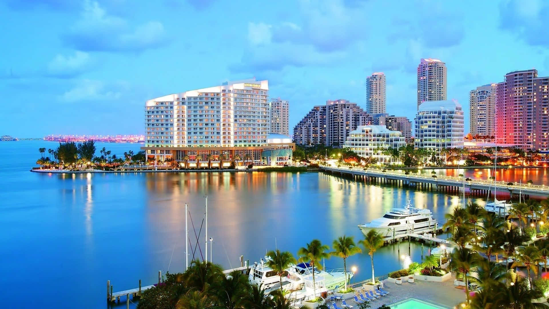 Miami 桌布 壁紙 — 黃昏時分充滿活力的海濱城市景觀，發光的摩天大樓倒映在平靜的水面上，周圍環繞著棕櫚樹和停泊的遊艇。