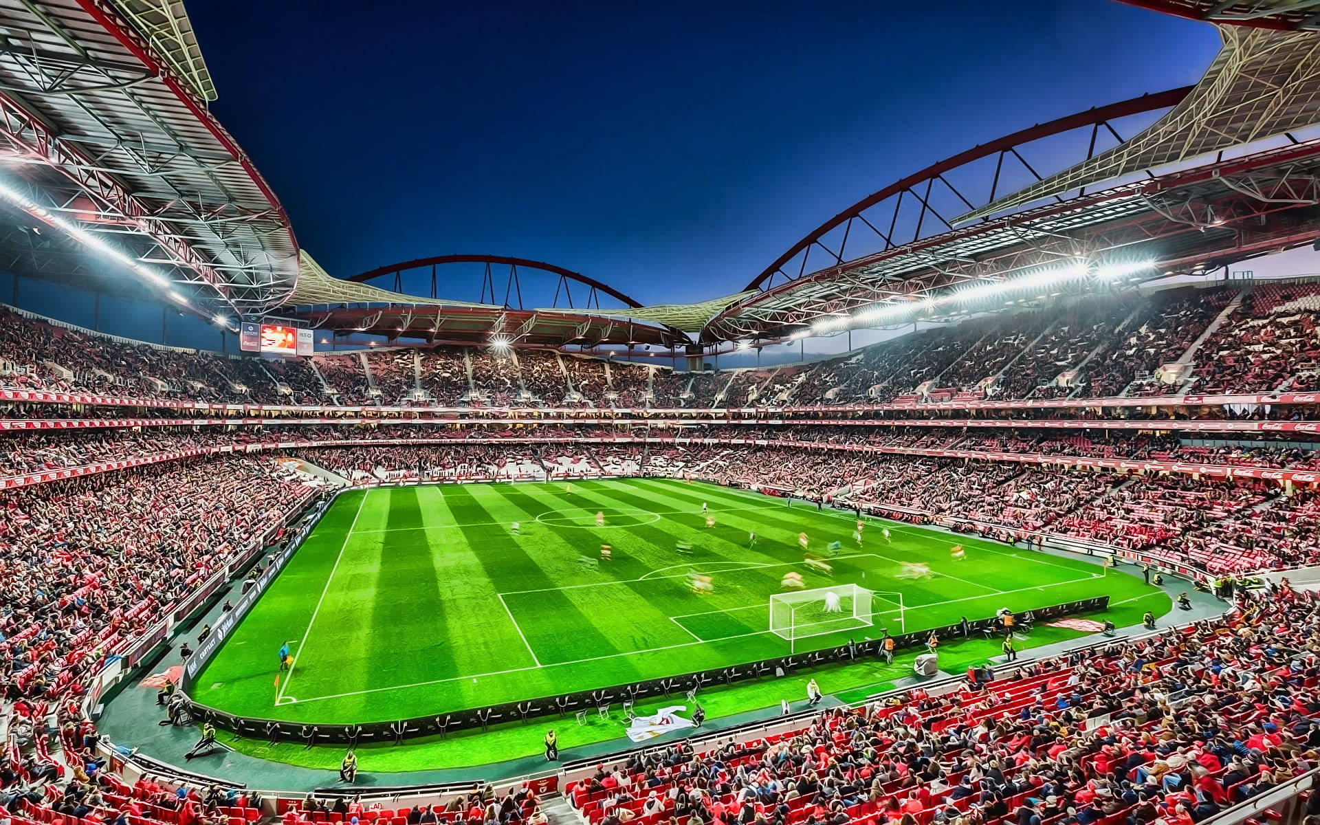 Stadium 桌布 壁紙 — 黃昏時分充滿活力的足球場，翠綠的草坪周圍是滿座的紅色座位，在閃爍的泛光燈下。