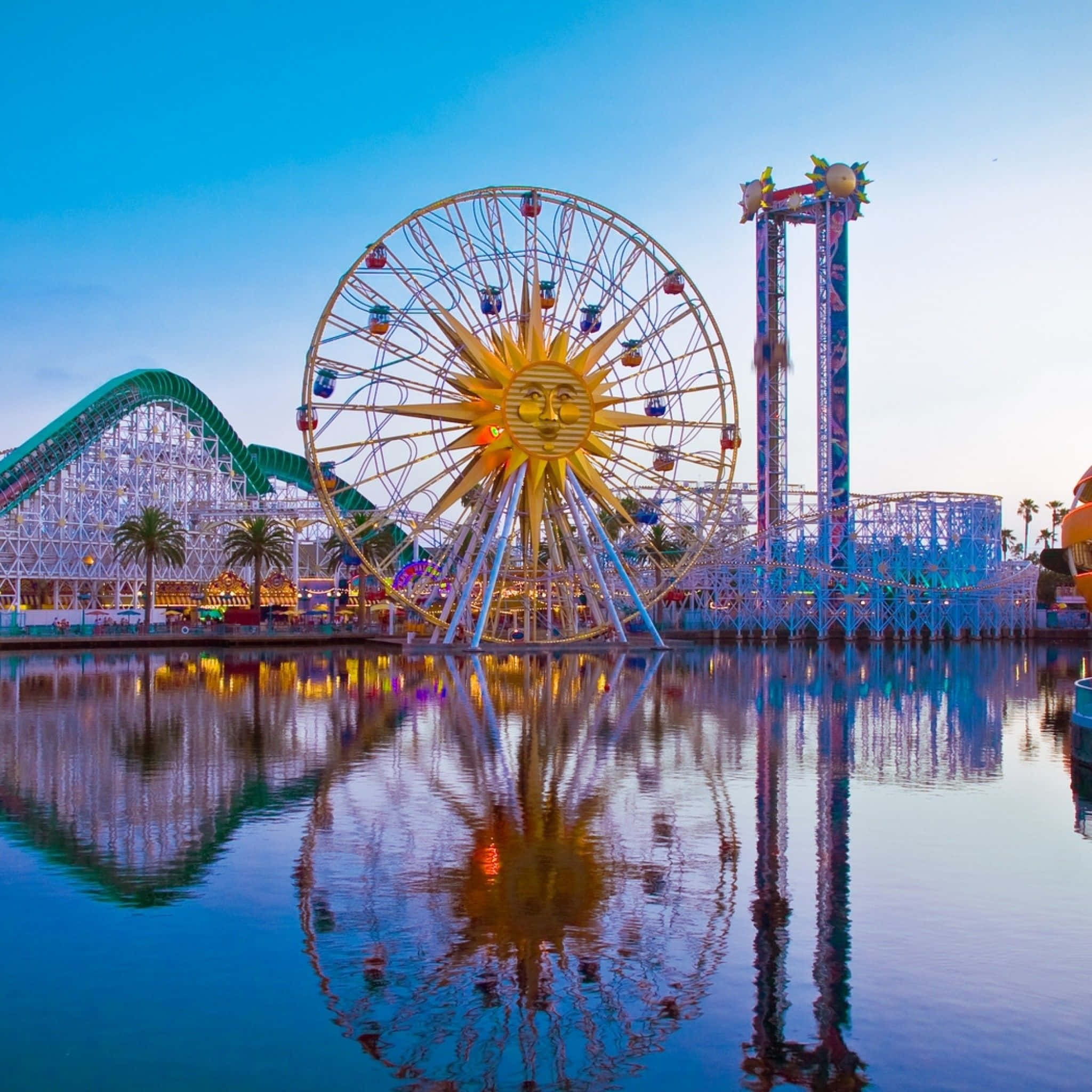 Disneyland 桌布 壁紙 — 黃昏時分充滿活力的遊樂園，太陽臉摩天輪和雲霄飛車倒映在平靜的水面上。