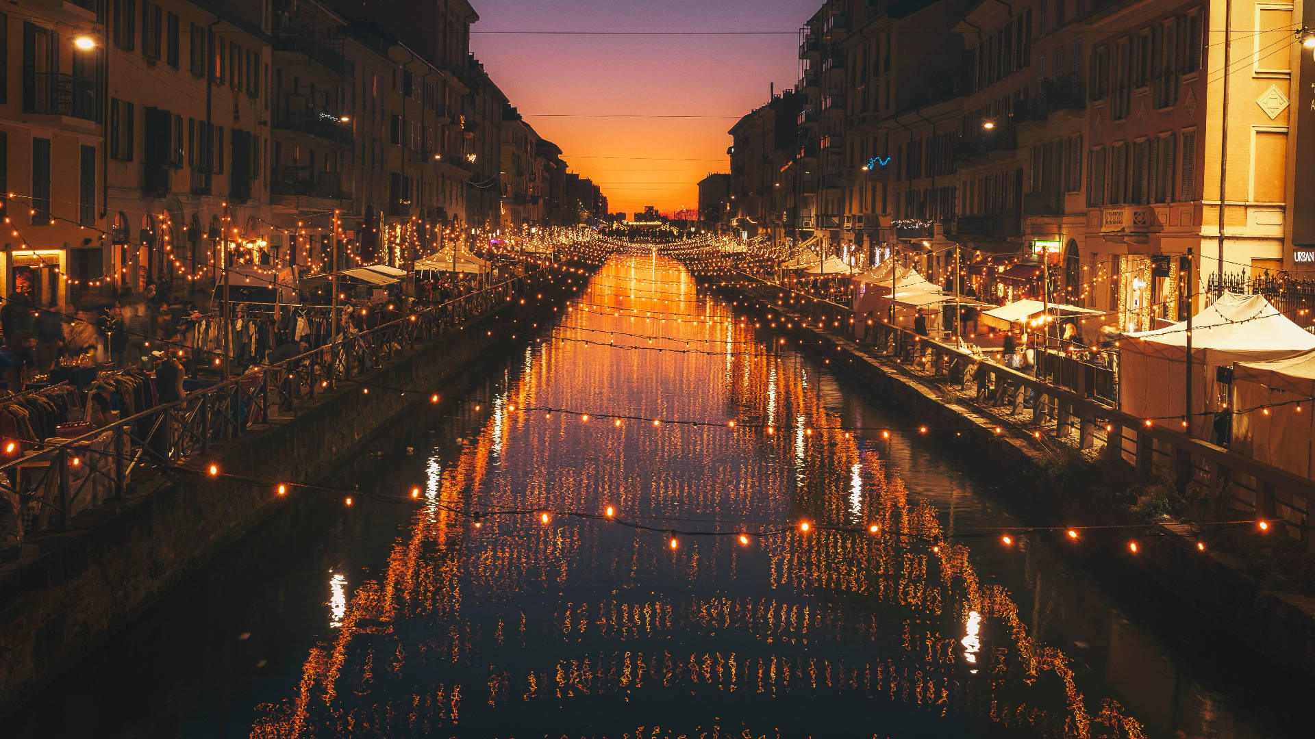 Milan 桌布 壁紙 — 黃昏時分充滿活力的運河，溫暖的串燈在水面上反射，兩旁是熙熙攘攘的咖啡館和歷史建築。