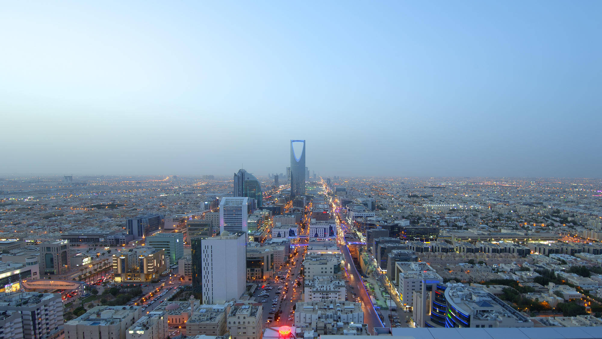 Riyadh 桌布 壁紙 — 黃昏時分利雅得天際線的鳥瞰圖，閃爍的路燈和標誌性的王國塔刺穿朦朧的天空。