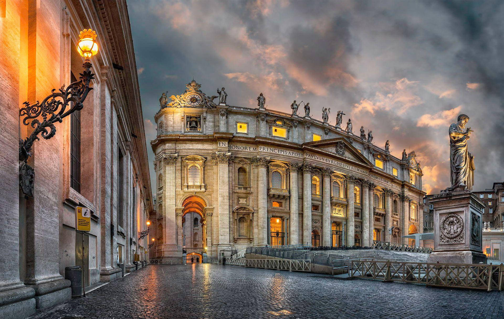Rome 桌布 壁紙 — 黃昏時分宏偉的聖彼得大教堂亮起燈光，濕漉漉的鵝卵石在戲劇性的雲層下反射出溫暖的光芒。