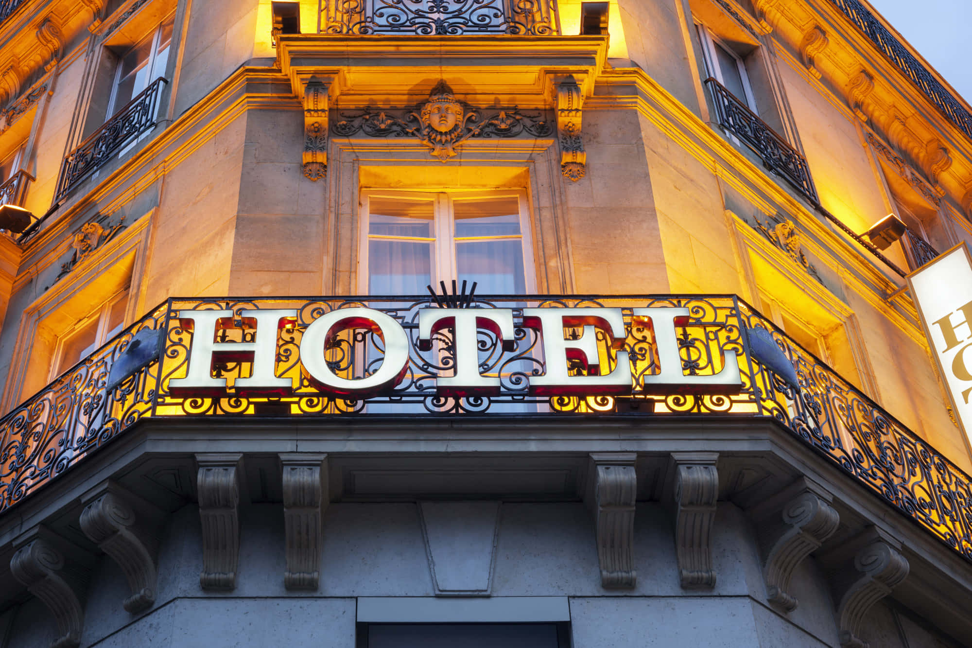 Hotel 桌布 壁紙 — 黃昏時分宏偉的飯店外牆，飾有華麗的鐵藝，發光的「HOTEL」招牌在石造建築上投下溫暖的金色光芒。