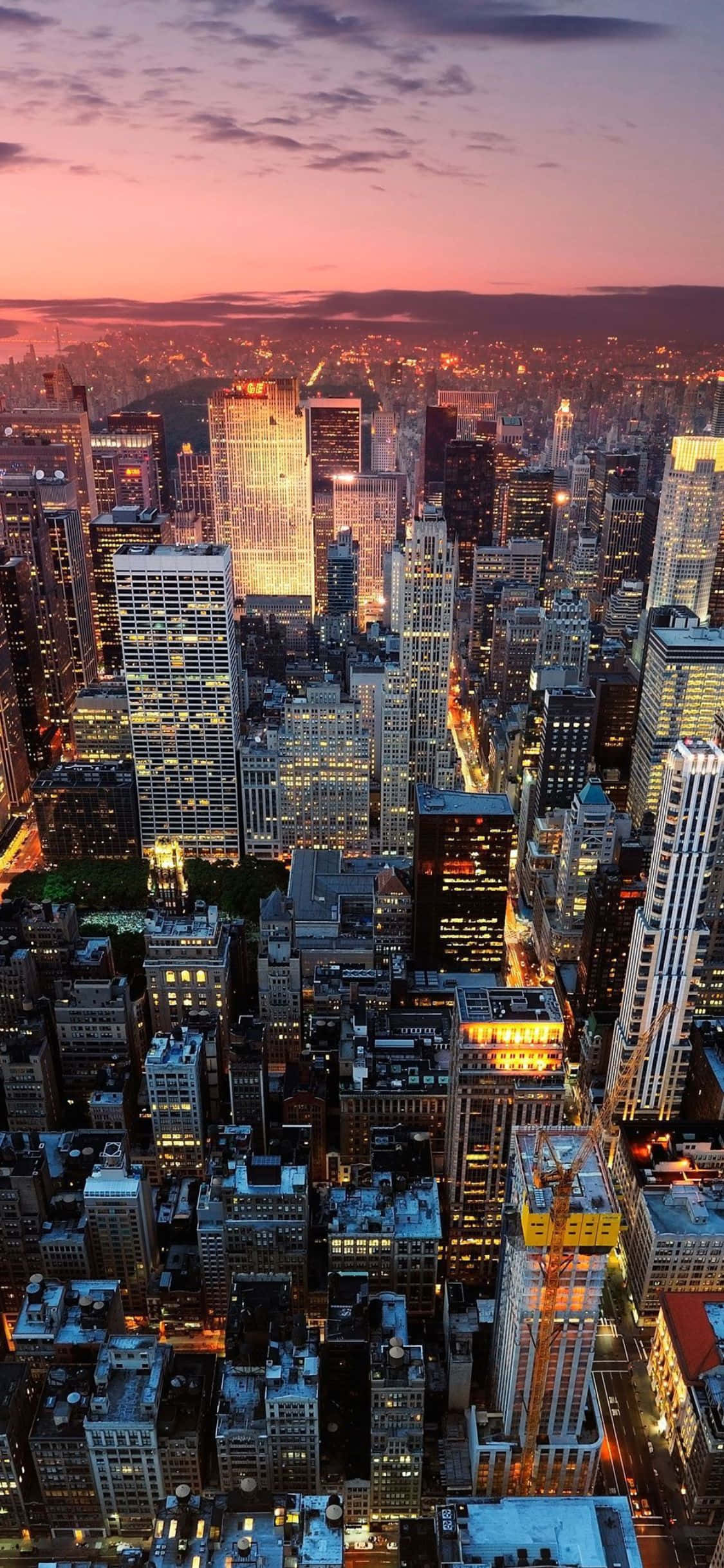 iPhone X New York 桌布 壁紙 — 黃昏時分密集城市景觀的鳥瞰圖，摩天大樓在粉紅色和紫色的天空下閃耀。