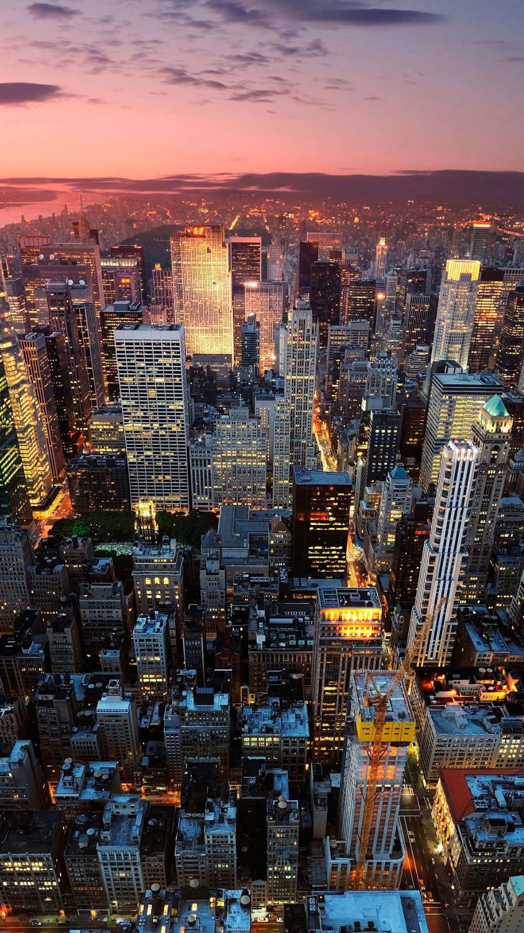 Android Ny City 桌布 壁紙 — 黃昏時分密集城市景觀的鳥瞰圖，摩天大樓在粉紅色和紫色的天空下閃閃發光。