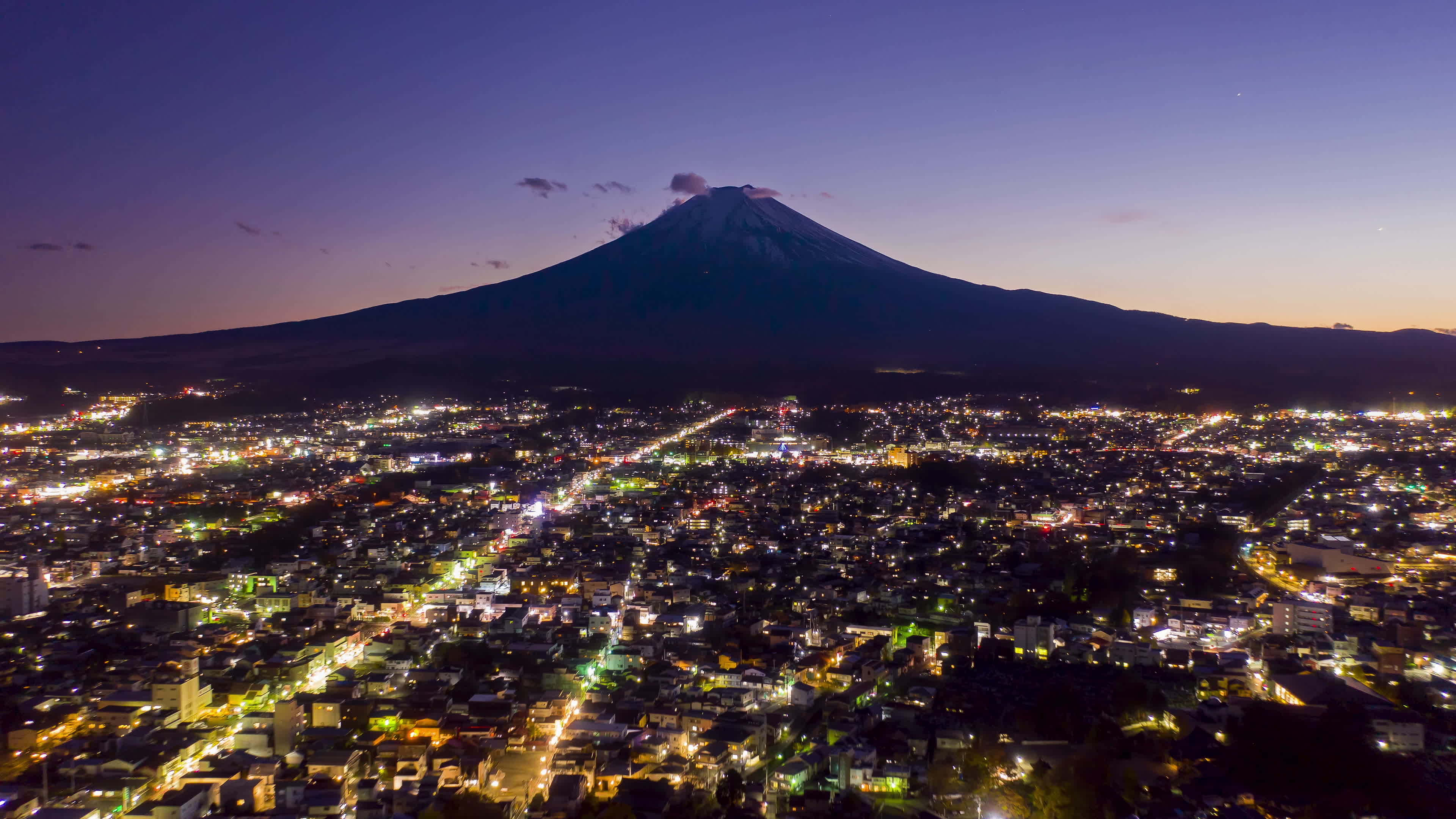 Mt Fuji 桌布 壁紙 — 黃昏時分富士山的鳥瞰圖，白雪覆蓋的山頂俯瞰著一座被溫暖路燈照亮的廣闊城市。