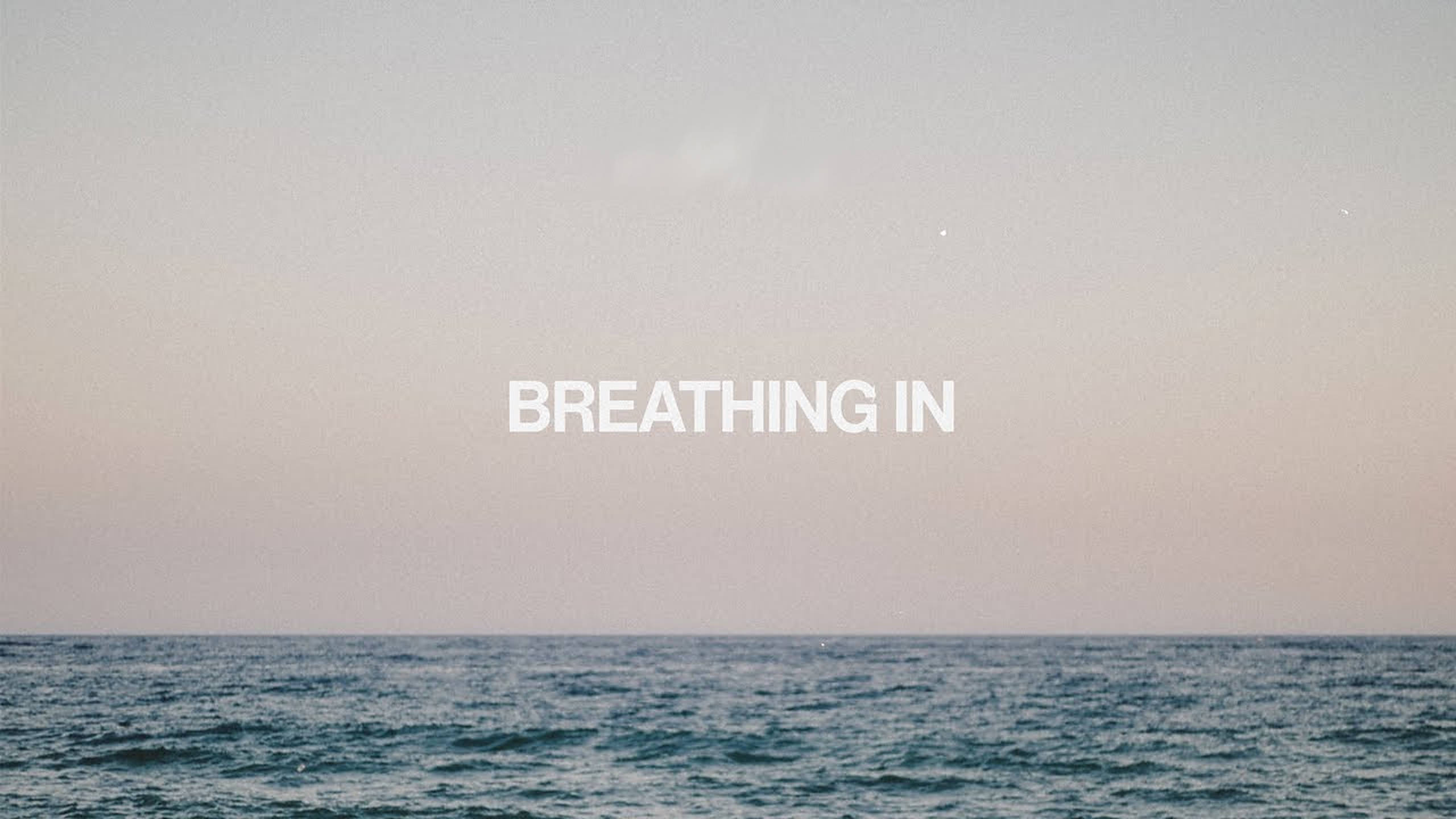 Breathing 桌布 壁紙 — 黃昏時分寧靜的海洋地平線，帶有柔和粉紅和藍色調，文字 breathing in 置於水面中央上方。