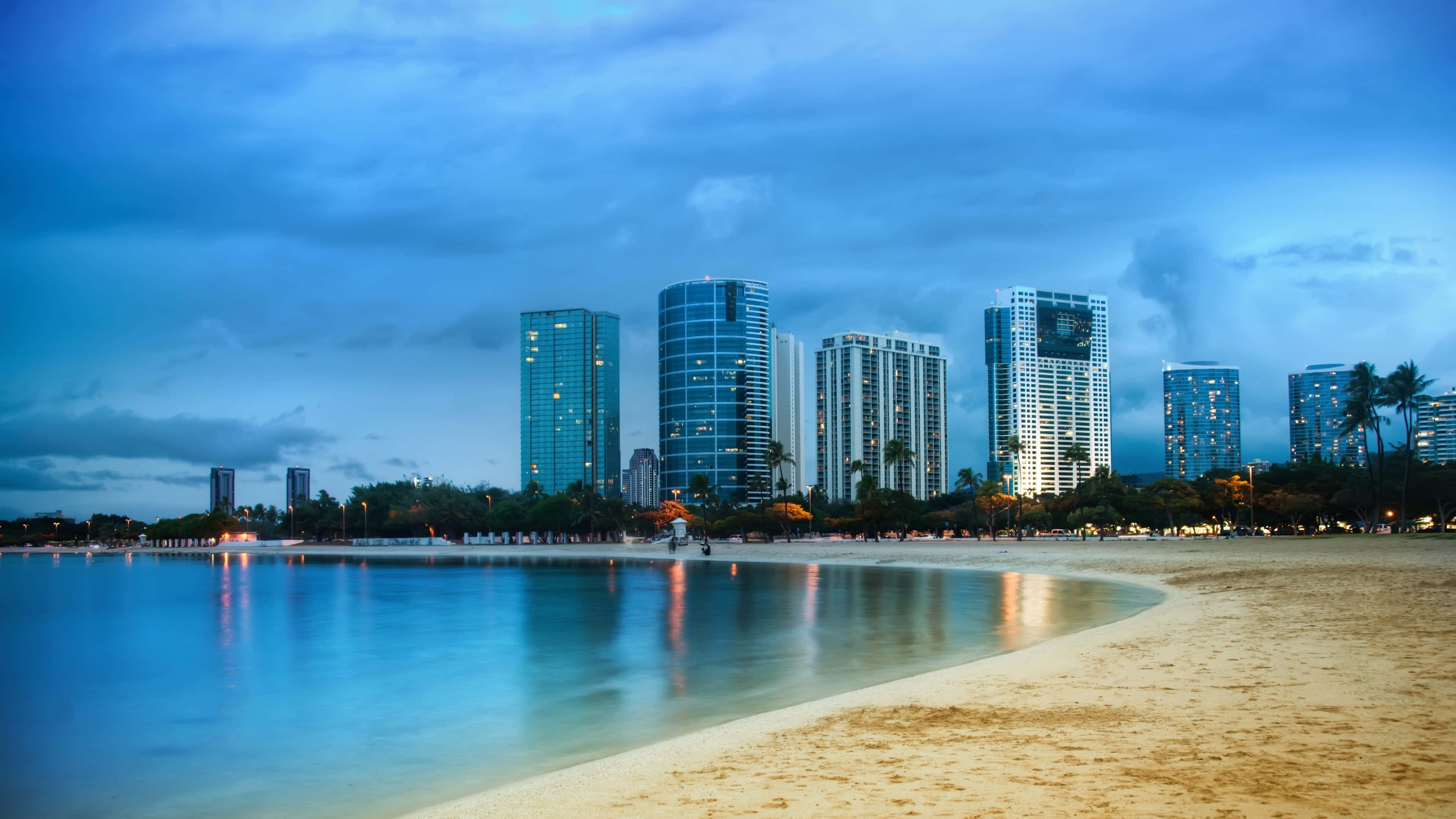 Miami 4K 桌布 壁紙 — 黃昏時分寧靜的海灘，平靜的水面倒映著高聳摩天大樓的城市燈光，天空呈現憂鬱的藍色。