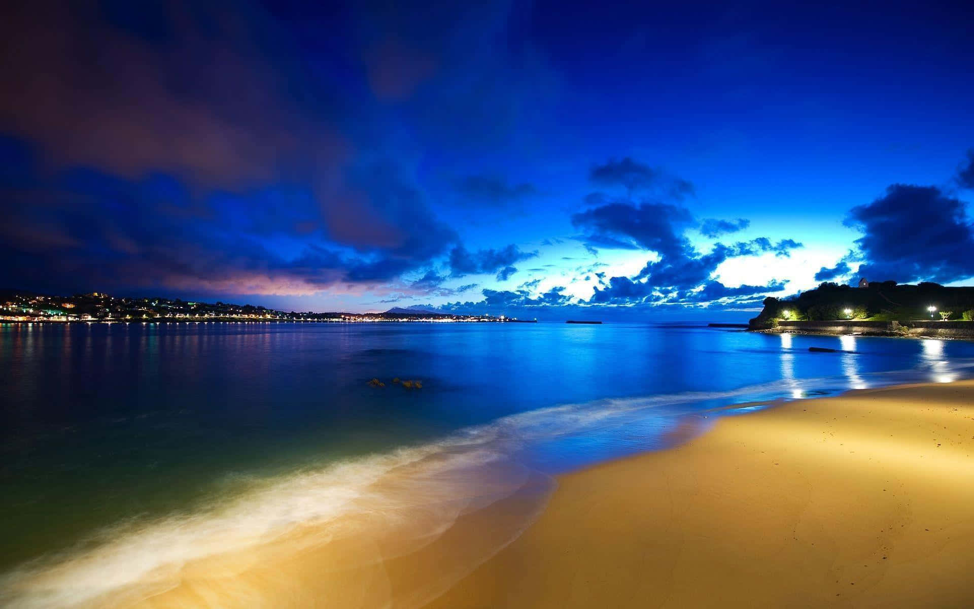 Beach Desktop 桌布 壁紙 — 黃昏時分寧靜的海灘，金色沙灘，平靜的藍色海水，遠處的城市燈光在戲劇性的多雲天空下反射。