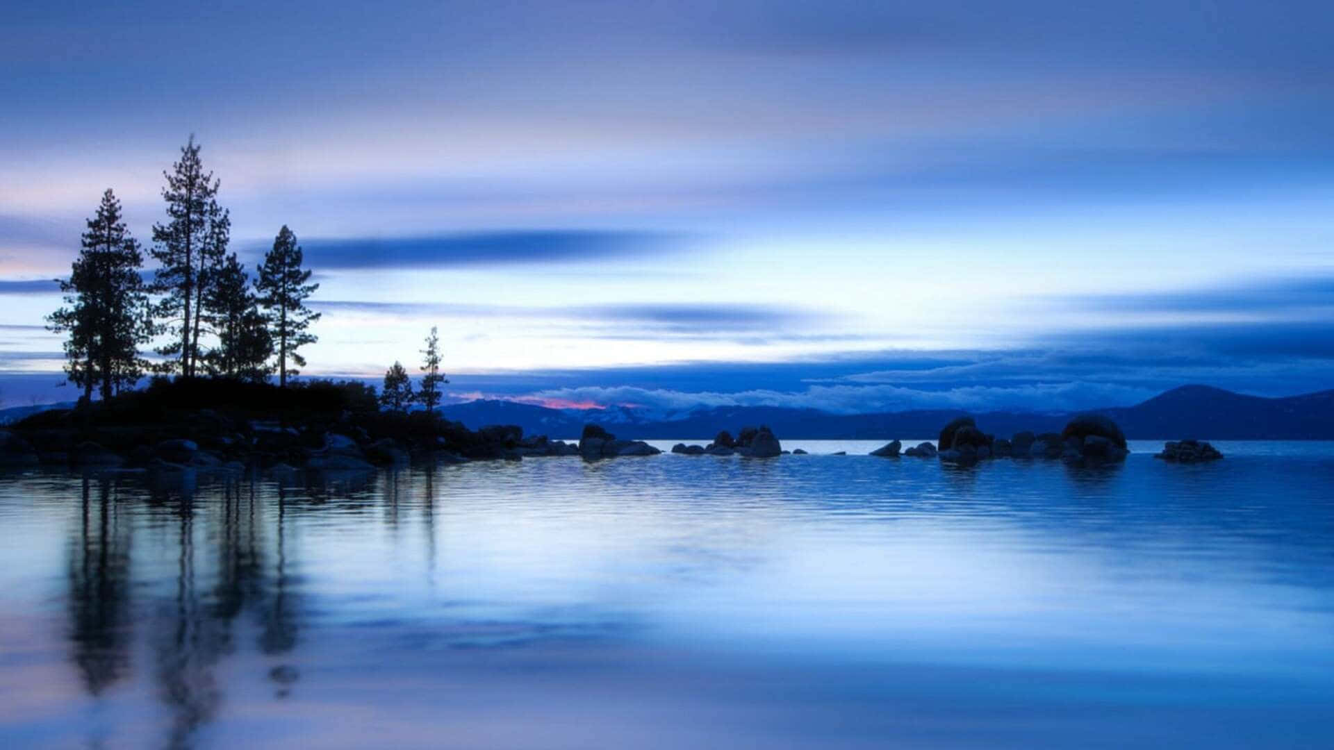 自然 桌布 壁紙 — 黃昏時分寧靜的湖景，岩石島上的樹木剪影在平靜的水面上倒映，天空柔和湛藍。